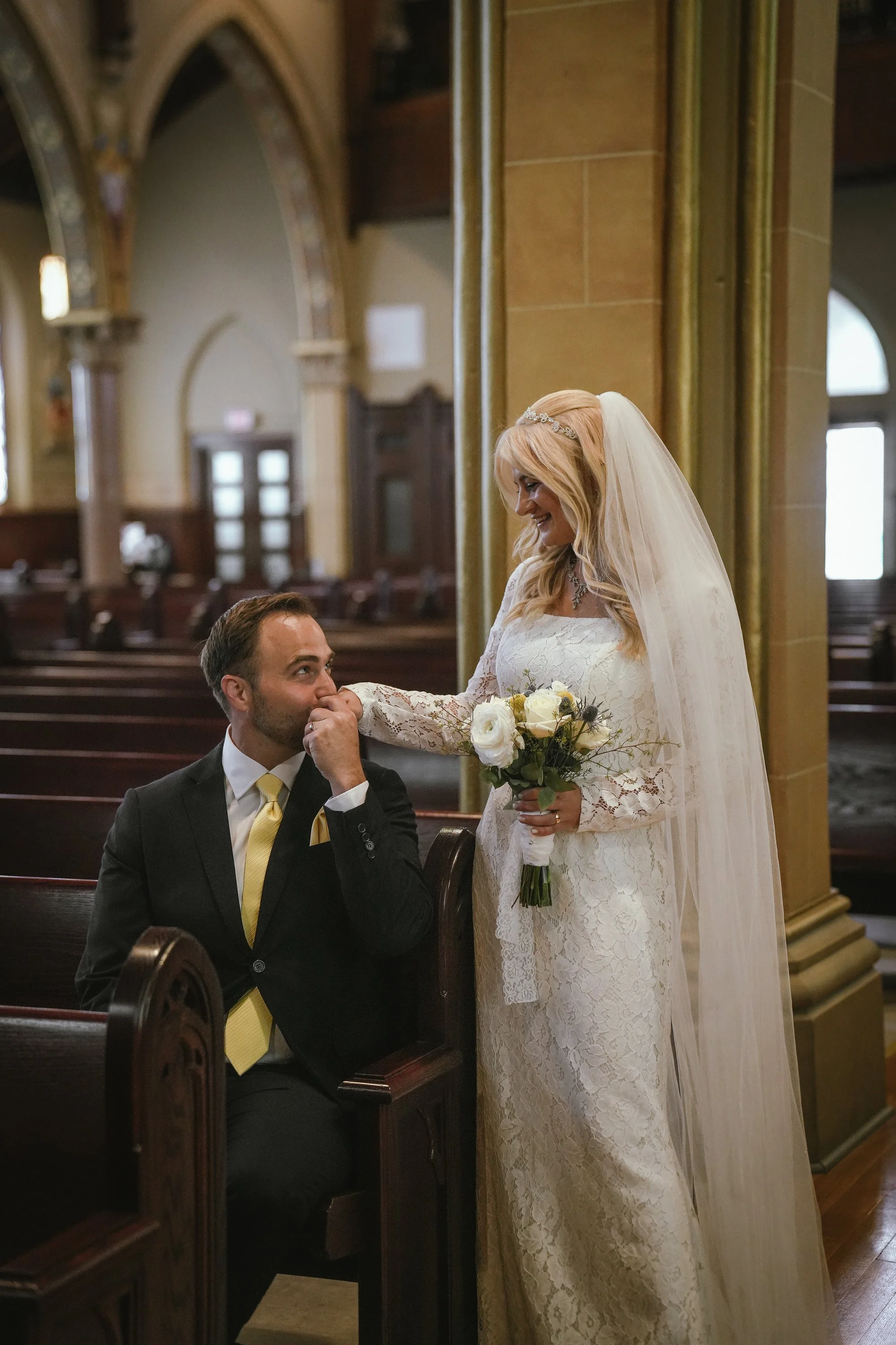 A bride in a lace wedding dress and veil holding a bouquet of white roses and greenery, standing in front of a groom. The groom, wearing a suit with a yellow tie, is seated in a church pew, looking up at the bride with a smile. The church interior features wooden pews and high arched ceilings.