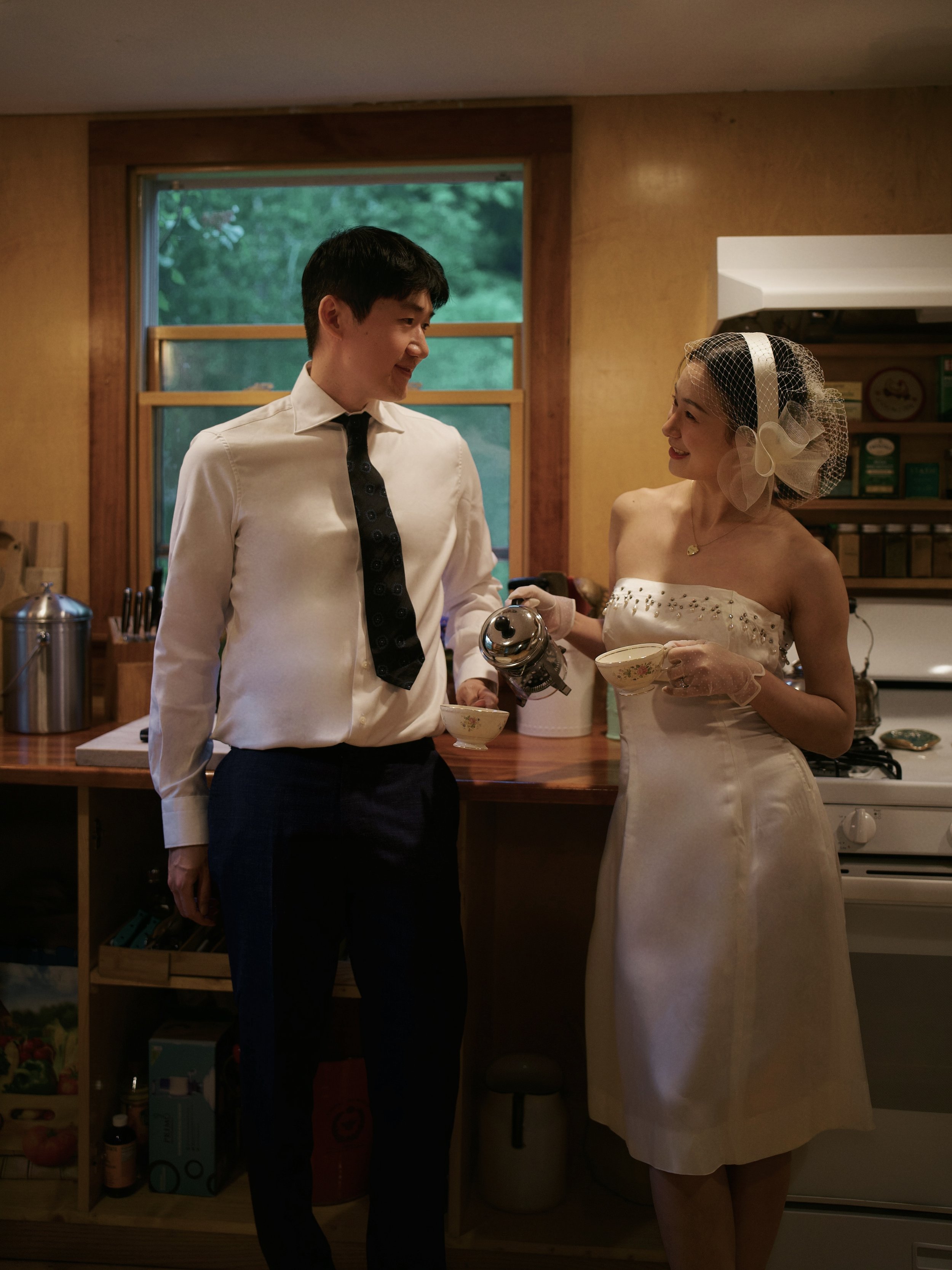 A couple in formal attire, with a man in a white shirt and black tie, and a woman in a white dress with a headpiece, standing in a kitchen. The woman is pouring a drink from a teapot into a teacup, while they smile at each other.