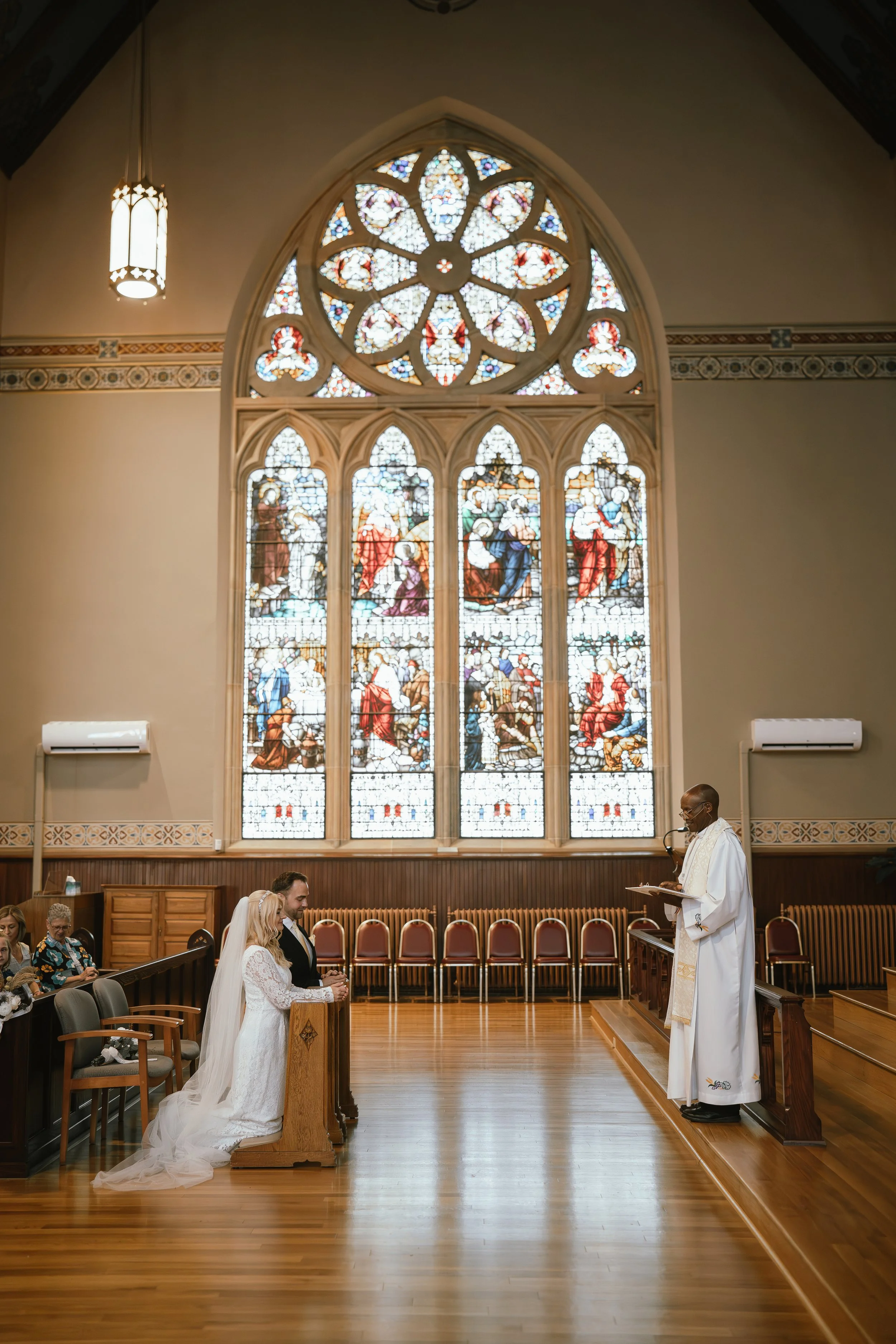 A wedding ceremony inside a church with stained glass windows depicting religious scenes. The bride and groom are kneeling at the altar, facing a priest. Pendant lighting, wooden pews, and decorative trim are visible.
