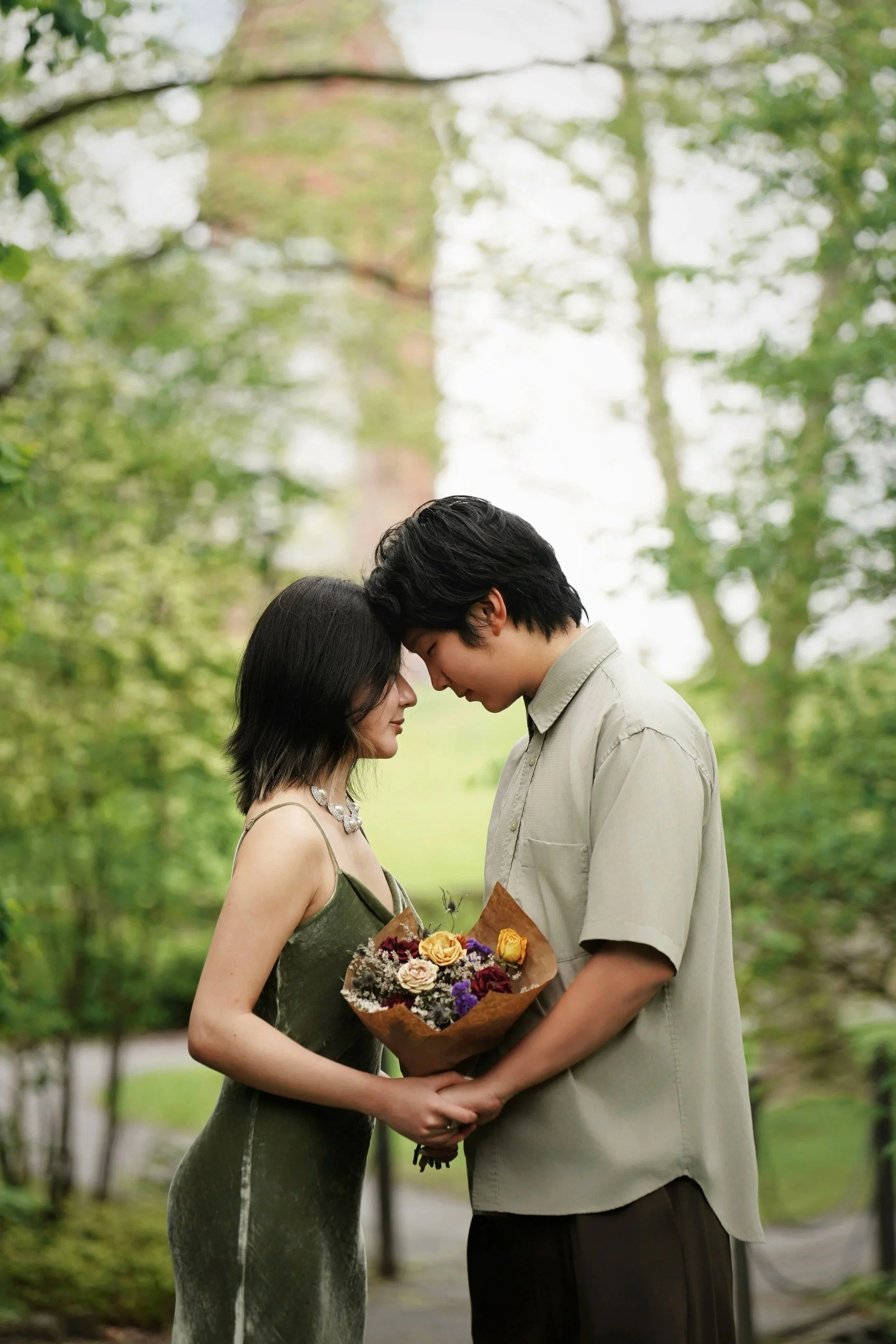 A young man and woman are standing close outdoors in a park, touching foreheads, with the woman holding a bouquet of flowers, both with gentle expressions, surrounded by trees.