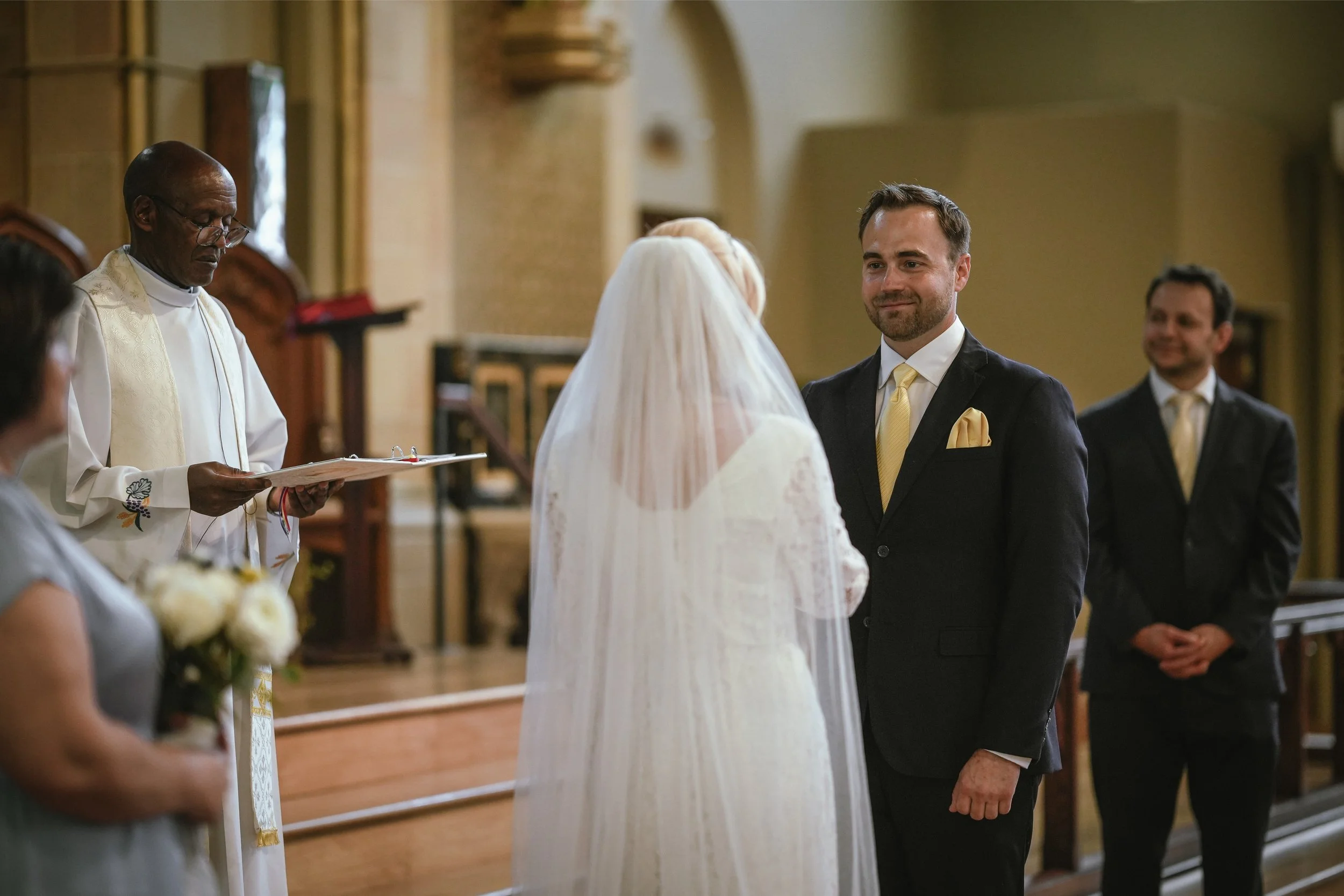 A groom in a dark suit with a yellow tie and pocket square smiling at his bride during a wedding ceremony inside a church, with a priest and a bridesmaid holding a bouquet in the background.