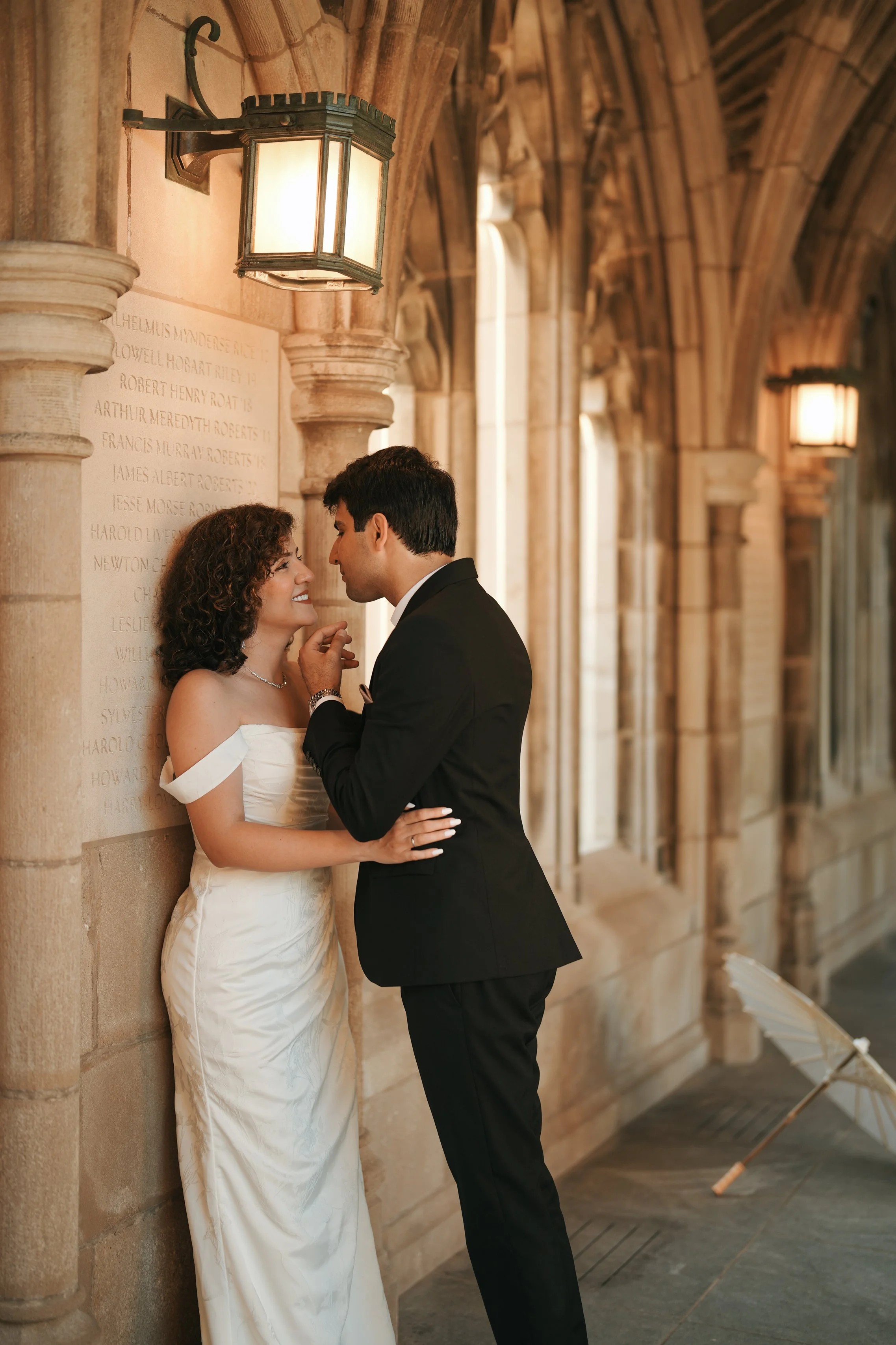 A woman in a white dress and a man in a black tuxedo stand close together in a historic hall with stone arches, with the woman smiling as they gently touch foreheads.