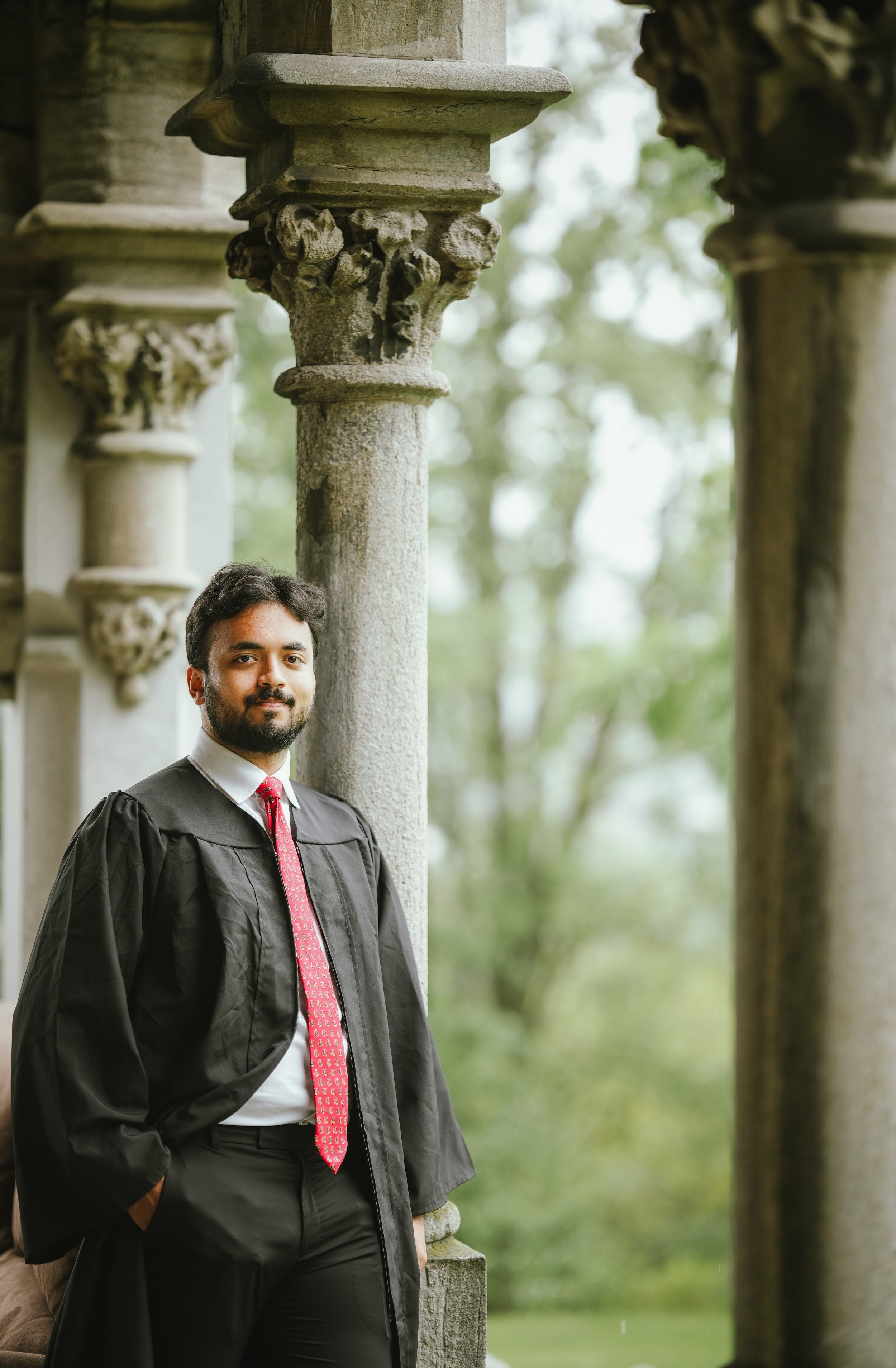 A man in a black graduation gown, white shirt, and red tie stands outdoors among stone columns and greenery, smiling slightly.