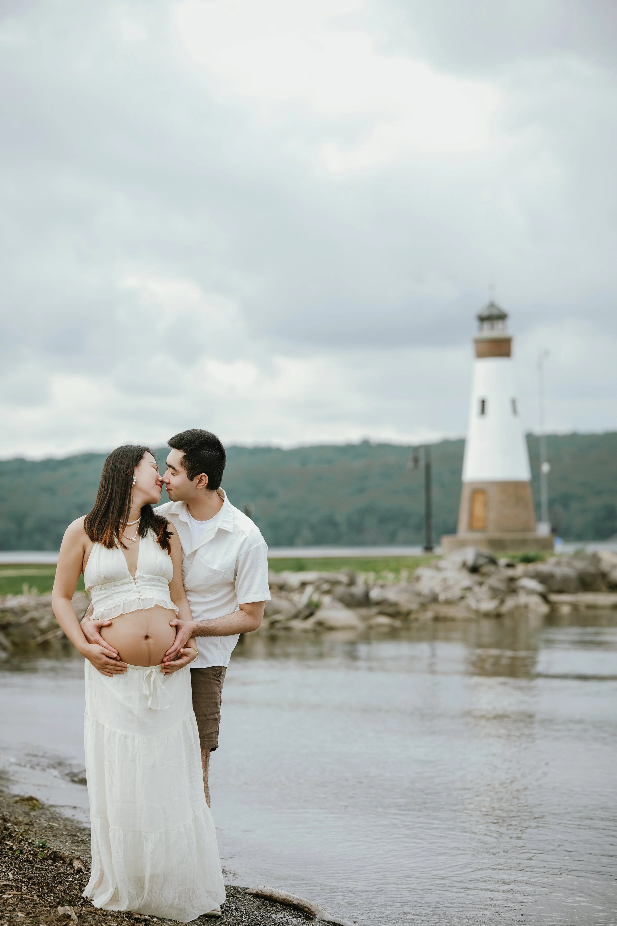 A pregnant woman and a man standing together near a river with a lighthouse and cloudy sky in the background. The man has his hands on the woman's pregnant belly, and they are touching noses.