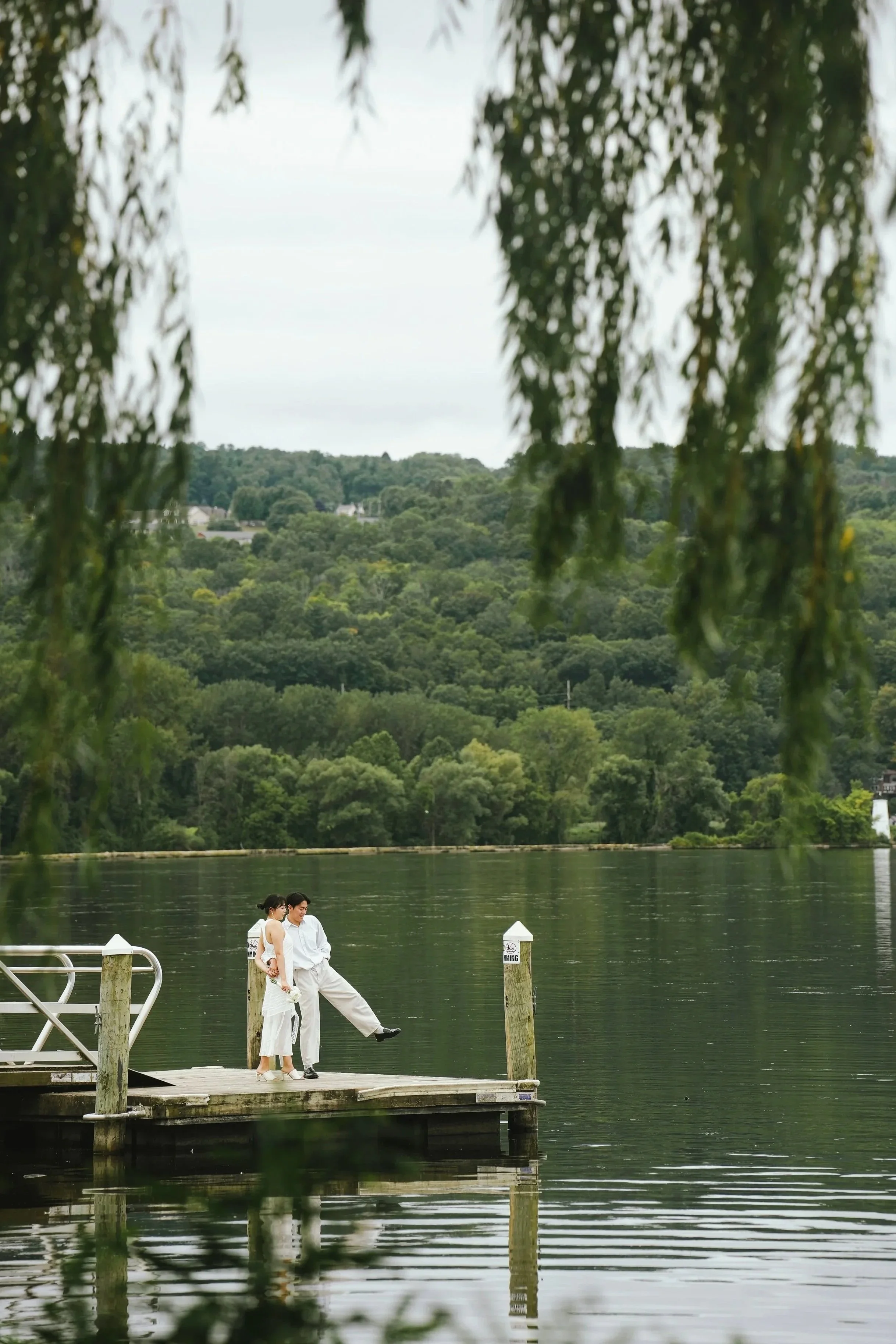 A couple dressed in white, standing on a dock by a lake with green hills in the background. The woman is wearing a white dress, and the man is dressed in white pants and a shirt. They are near a white railing, with a tree with hanging leaves partially obscuring the view.