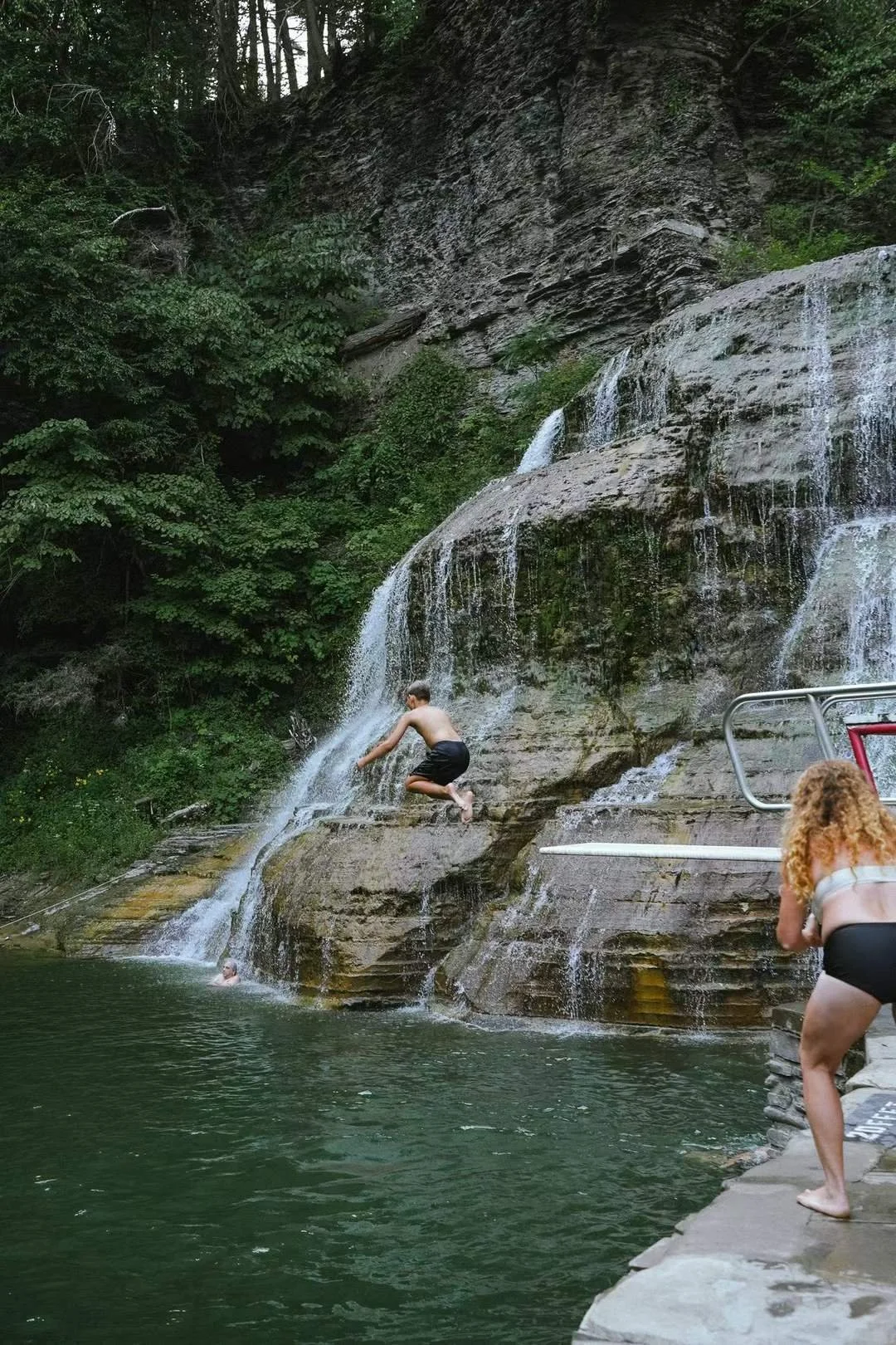 Children playing and jumping into a natural swimming hole beneath a waterfall surrounded by forested rocks and trees.