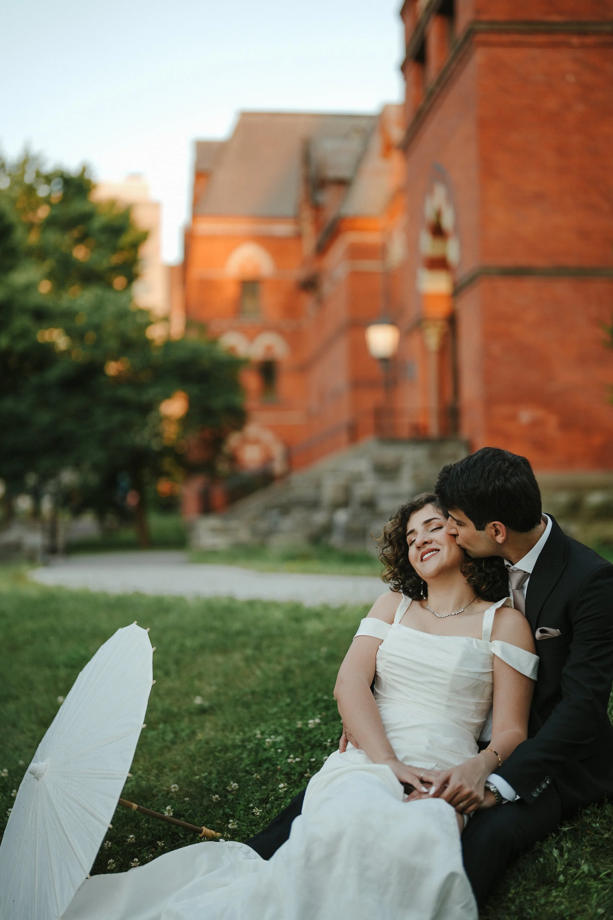 A couple dressed in wedding attire sitting on the grass, with the groom kissing the bride on the cheek. There is a white umbrella on the ground nearby in a park with a large brick building in the background.