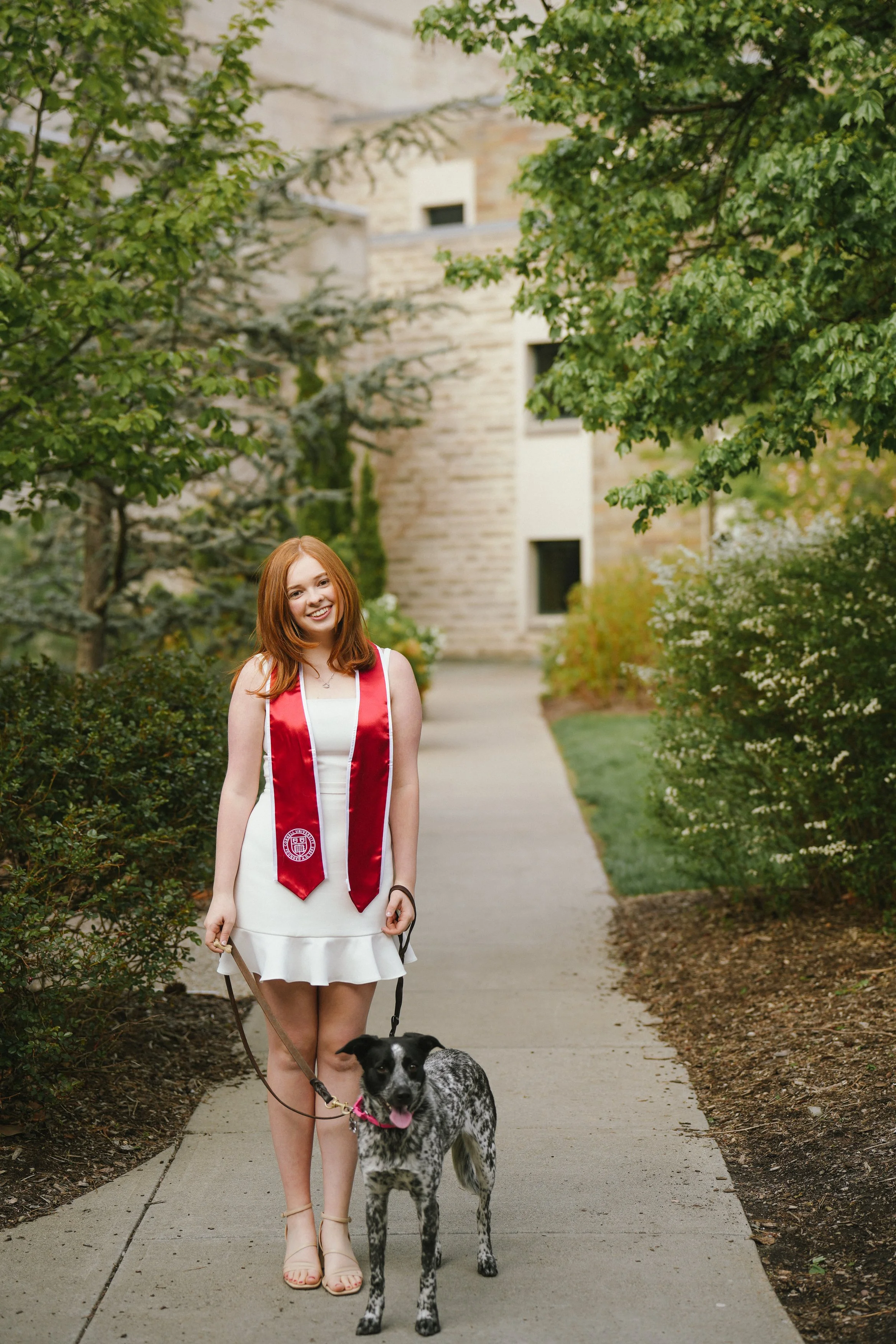 Young woman in a white dress and beige heels holding a leash with a black and white dog, standing on a sidewalk surrounded by greenery and trees.