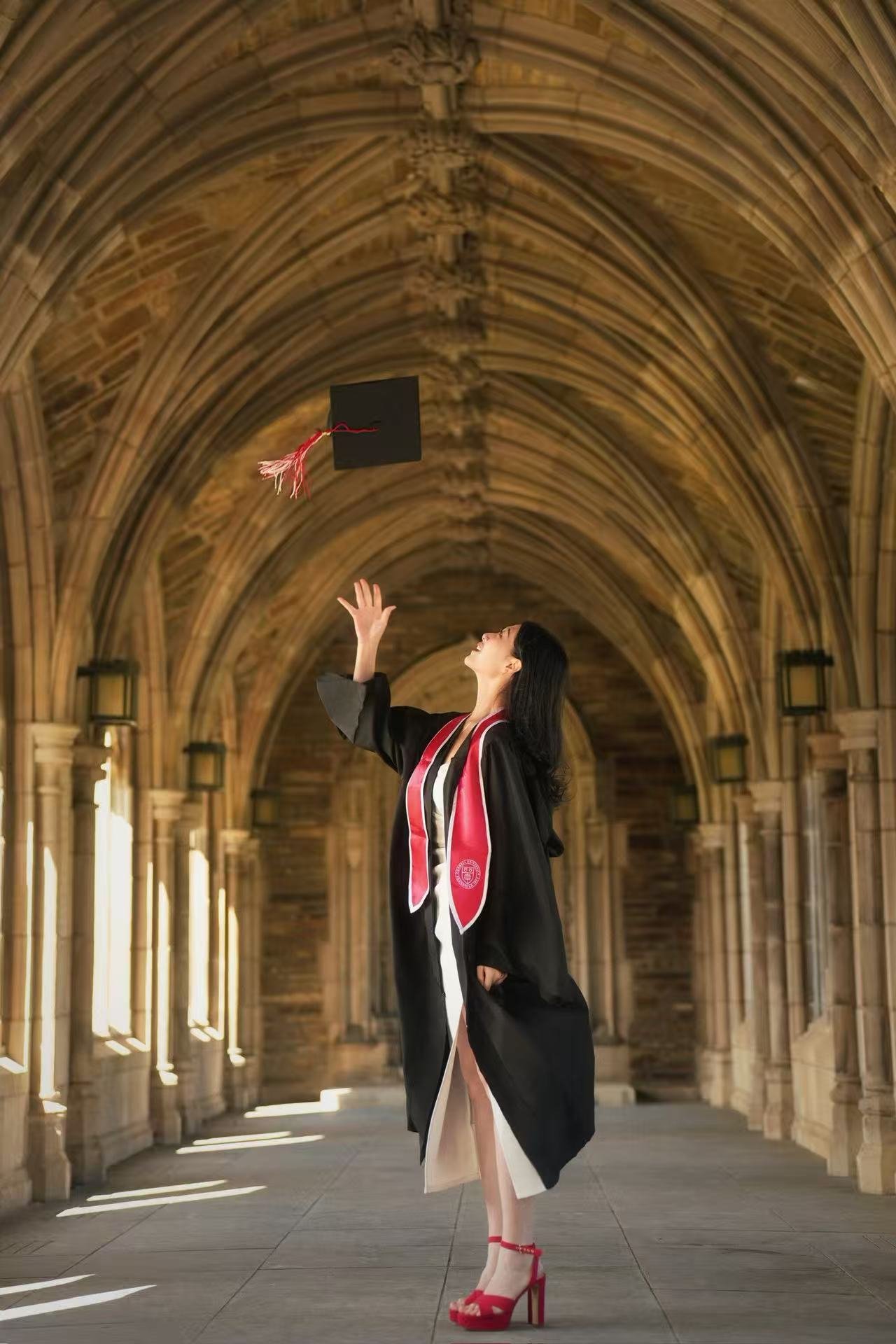 A woman in a graduation gown and stole celebrating inside a gothic-style corridor with arched ceilings, throwing her graduation cap in the air.