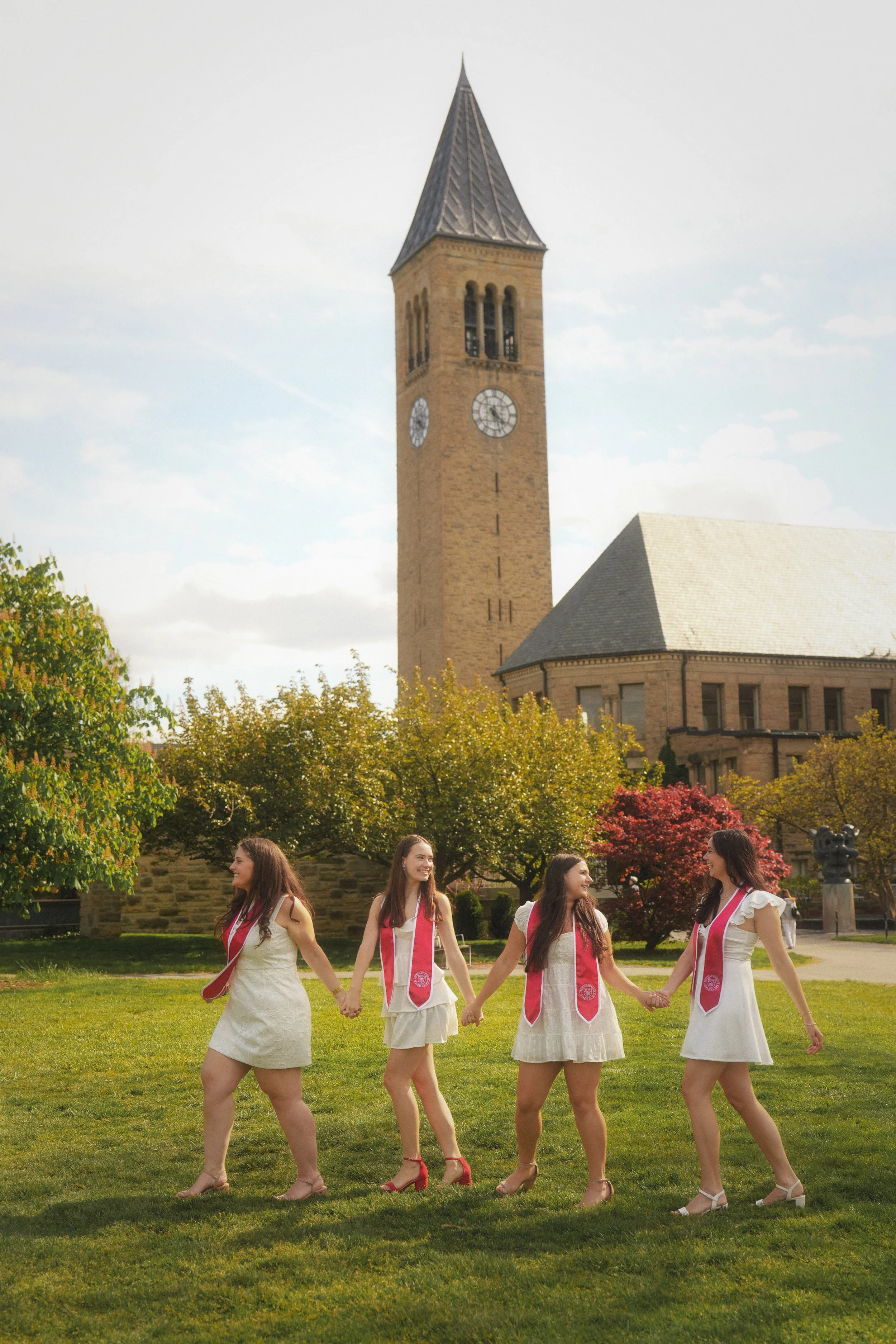 Group of five young women in white dresses with pink sashes, holding hands and dancing outdoors in a park with green grass, trees, and a historic clock tower in the background.