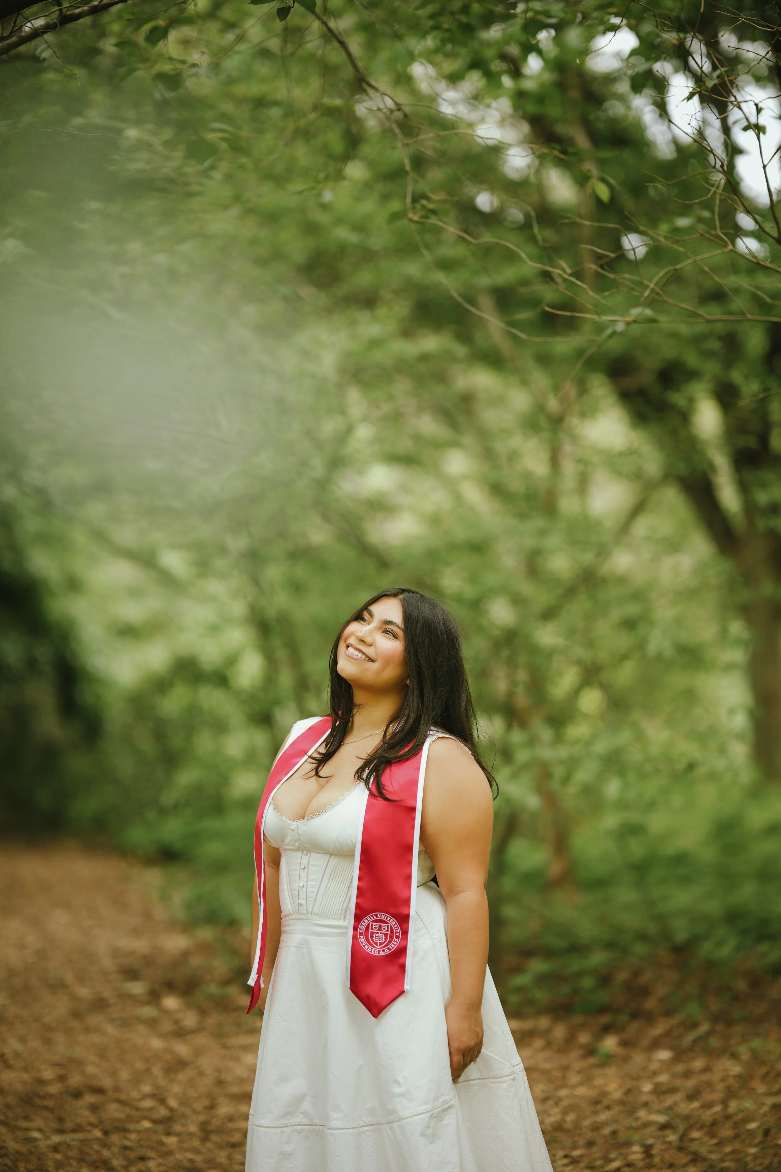 A young woman in a white dress with a red graduation stole, standing outdoors in a wooded area, smiling with her head slightly tilted.