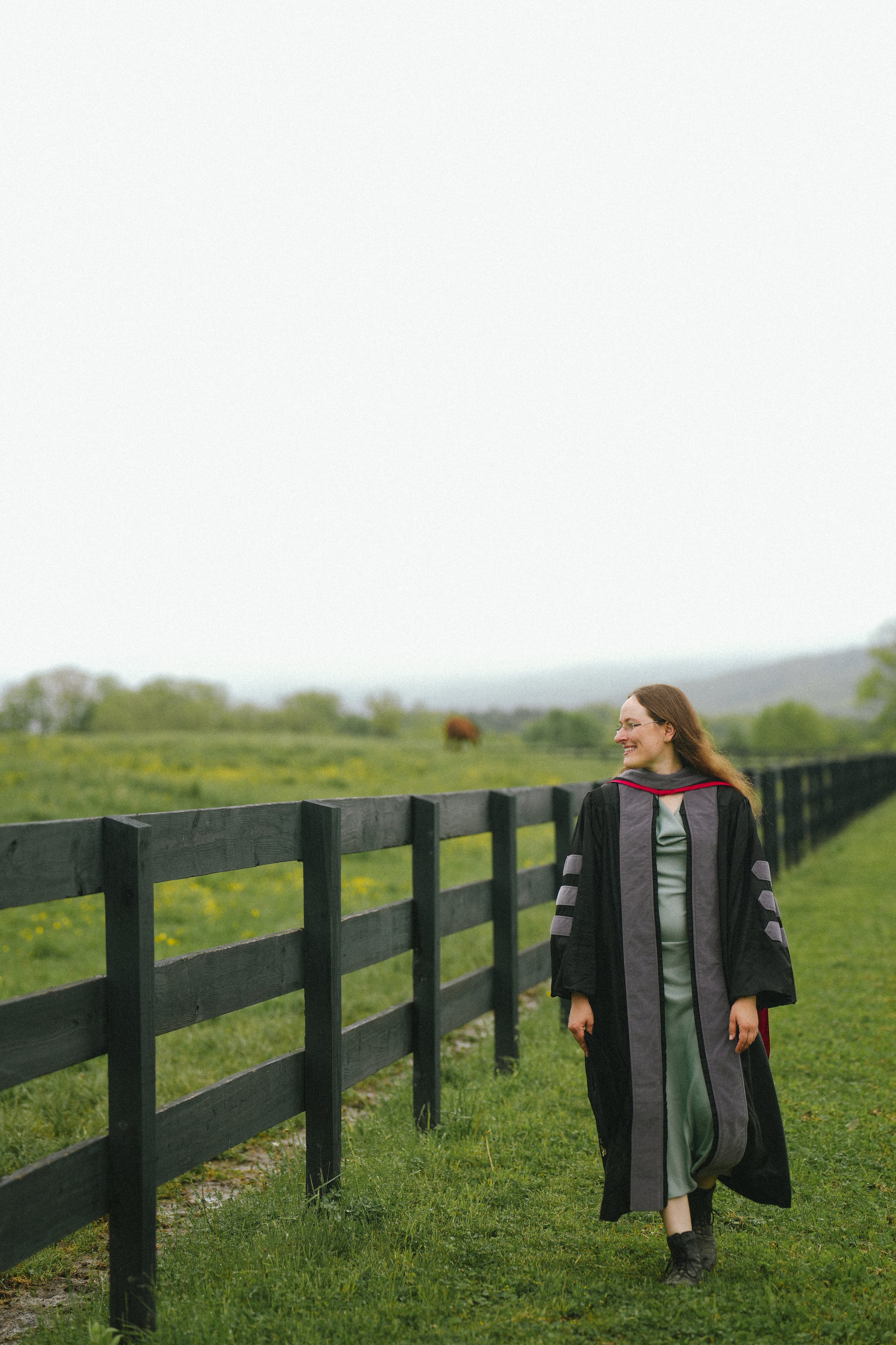 A woman in graduation regalia walking outdoors on grass, with a black fence and cloudy sky in the background.