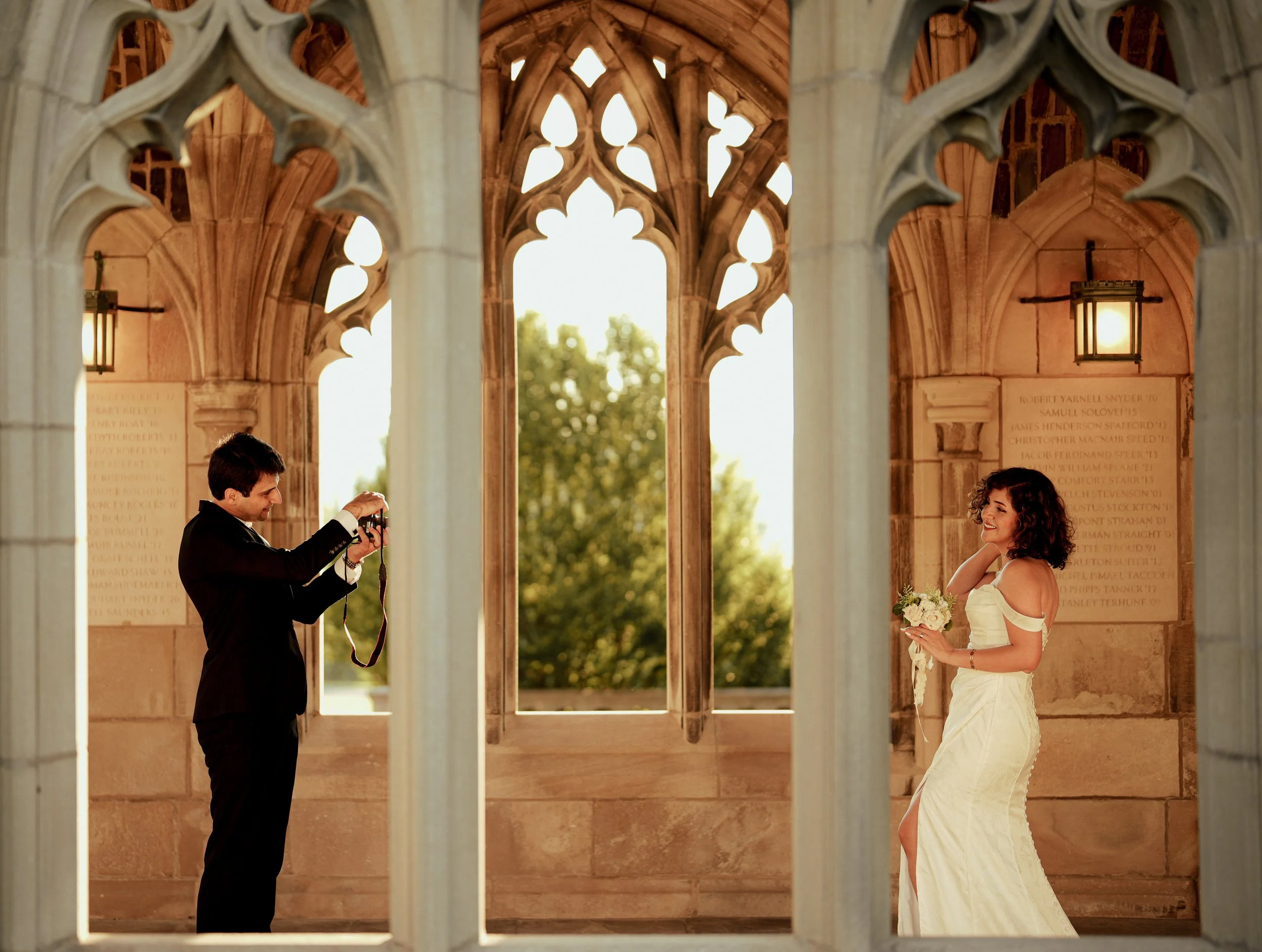 A man in a tuxedo taking a picture of a woman in a white wedding dress holding a bouquet of flowers in a historic stone architectural setting with arches and hanging lanterns.
