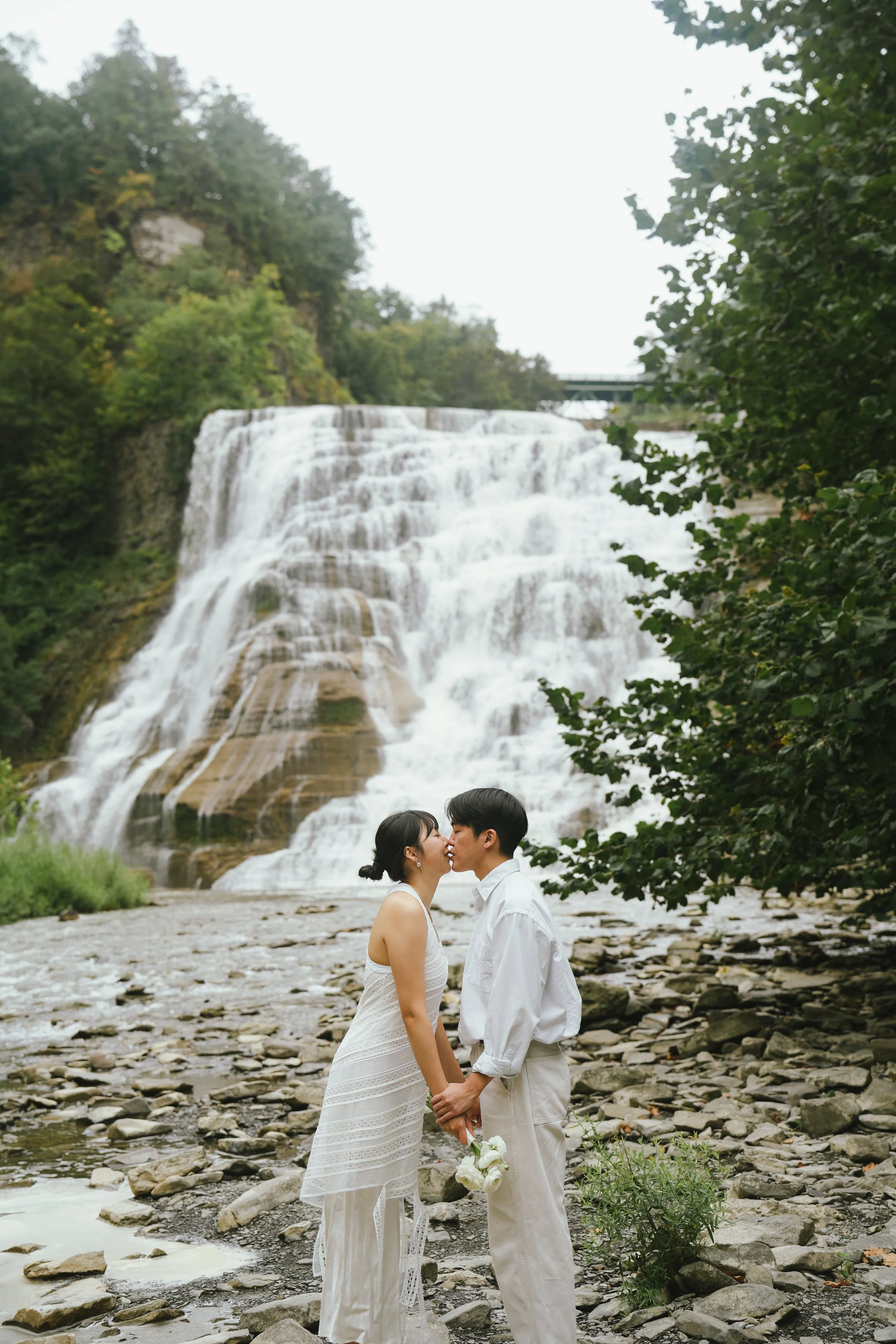 A couple dressed in white, holding hands and about to kiss, stands on a rocky riverbank with a large waterfall and trees in the background.