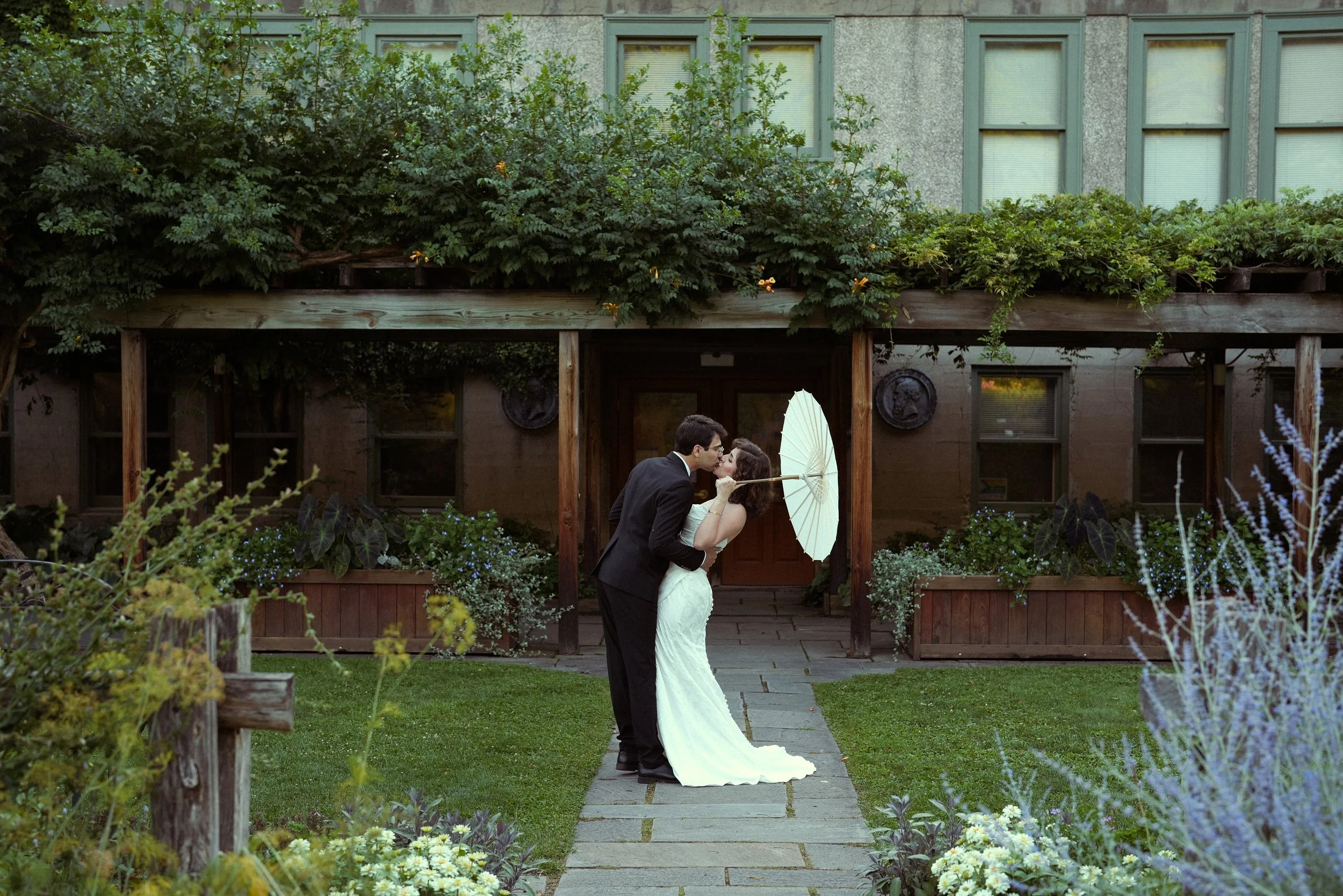 A bride and groom kiss outdoors in front of a house with large windows, surrounded by greenery and flowers.