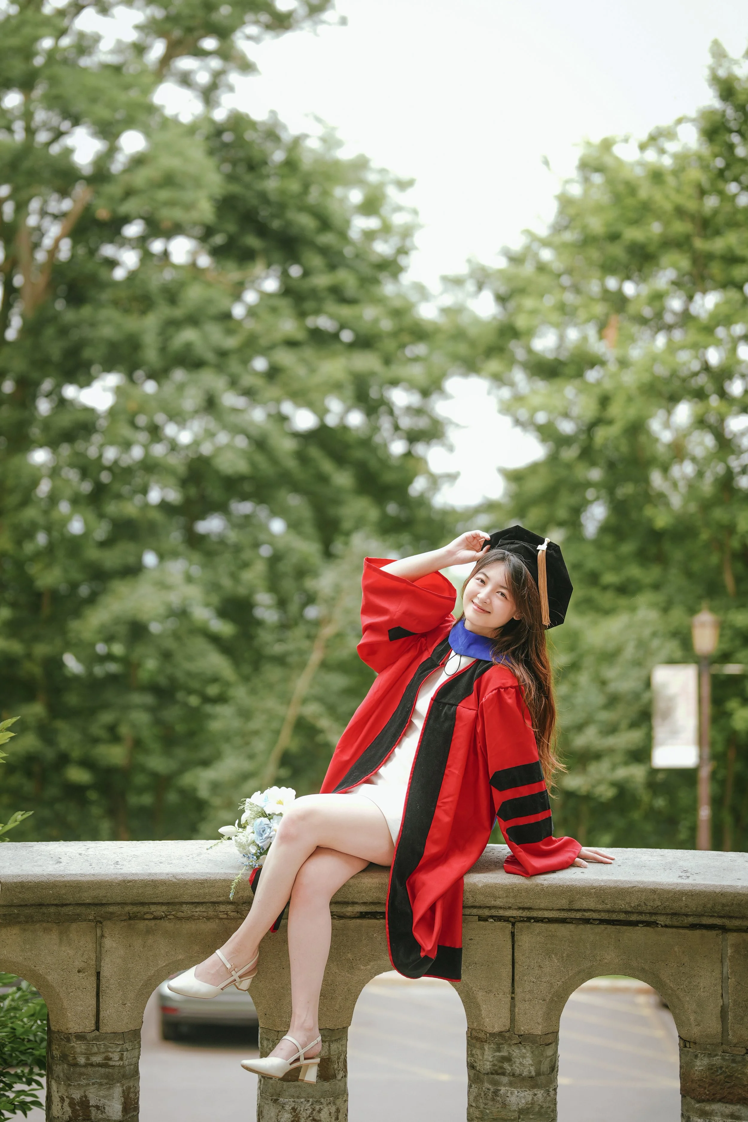 A young woman wearing a red graduation gown and cap, sitting on a stone balustrade outdoors in a park with trees in the background, smiling and posing for a photo.