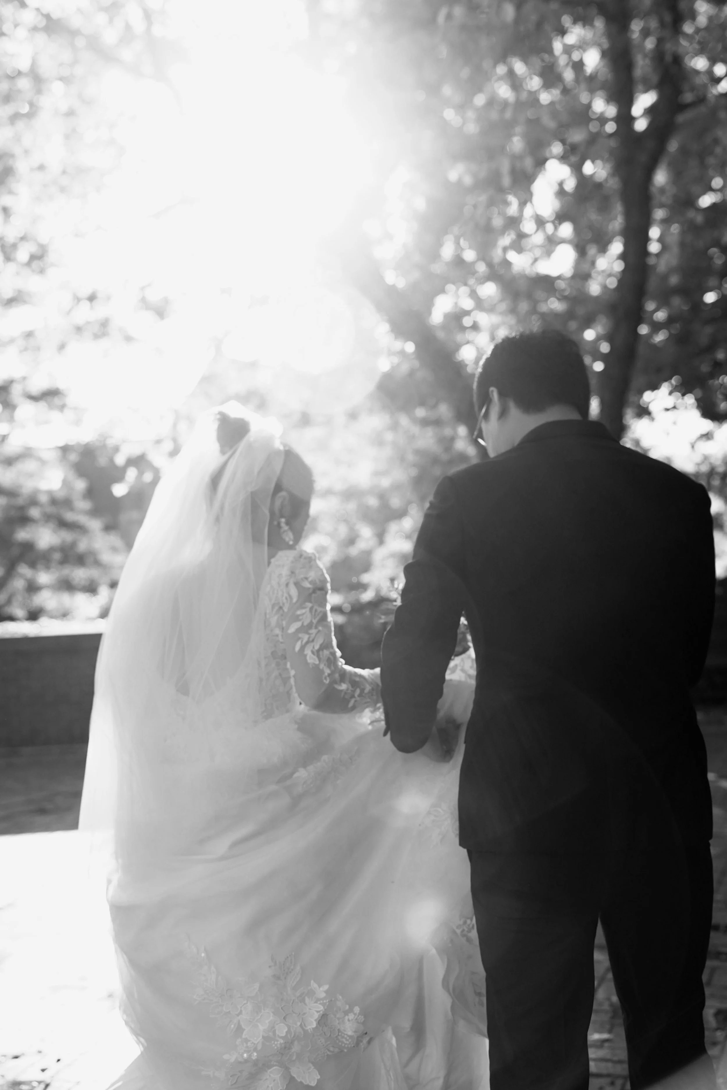 A black and white photo of a bride and groom holding hands outdoors, with the sun shining through the trees in the background.
