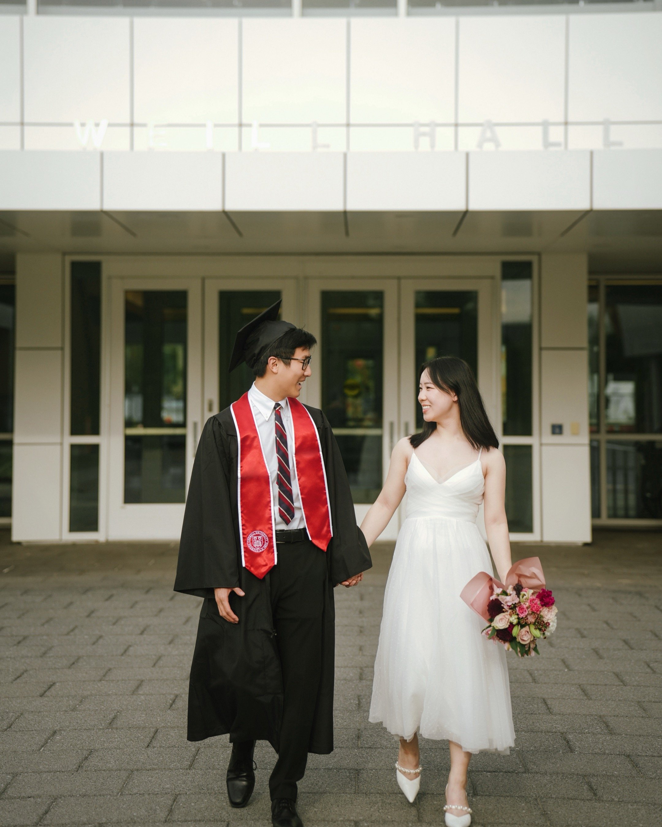 A happy couple, a groom in a traditional black graduation gown and cap, and a bride in a white dress, holding hands and smiling at each other outside a building with glass doors. The bride is holding a bouquet of flowers.