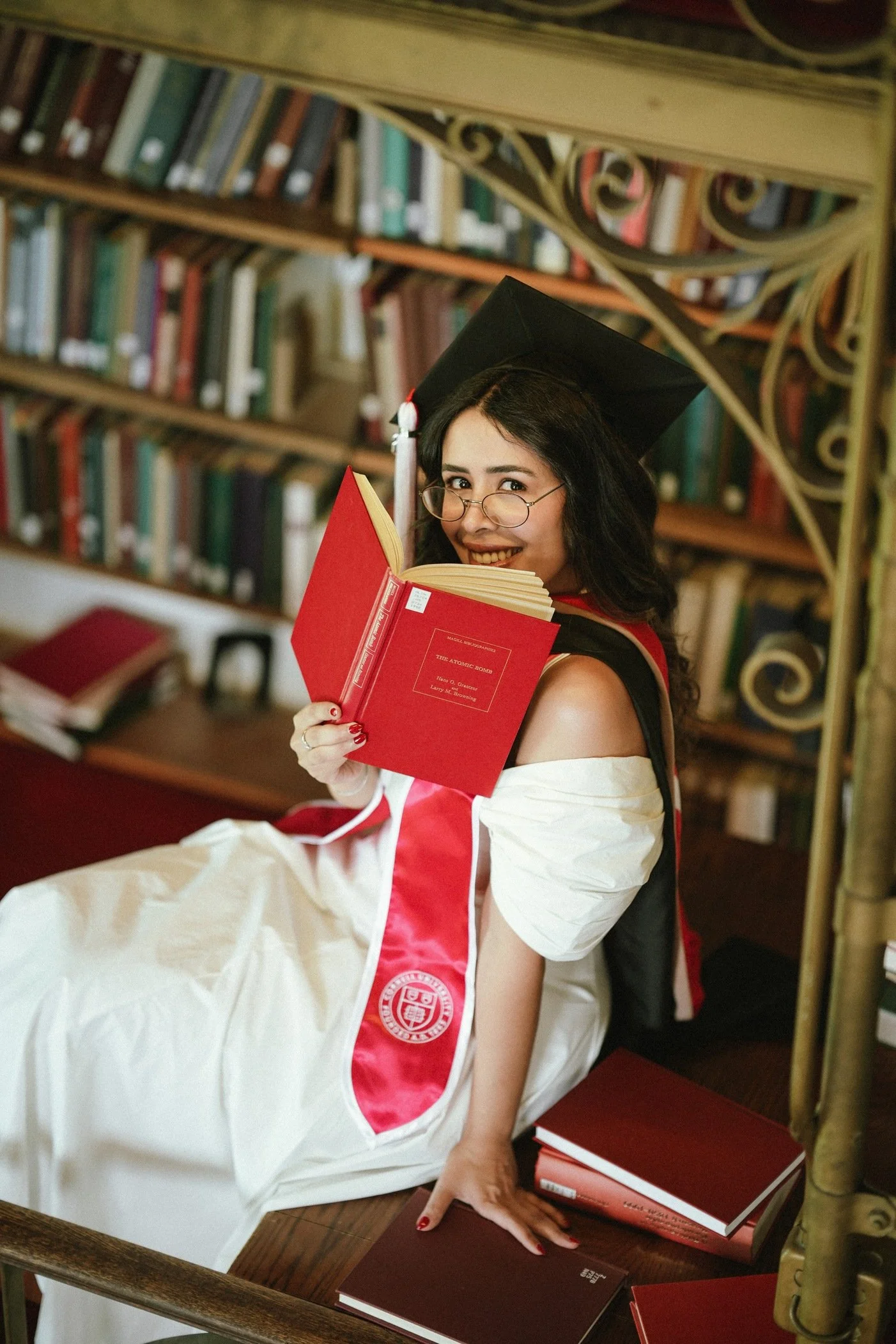 A young woman dressed in graduation attire, including a cap and gown, is smiling and looking over her shoulder while holding an open red book in a library with shelves filled with books behind her.