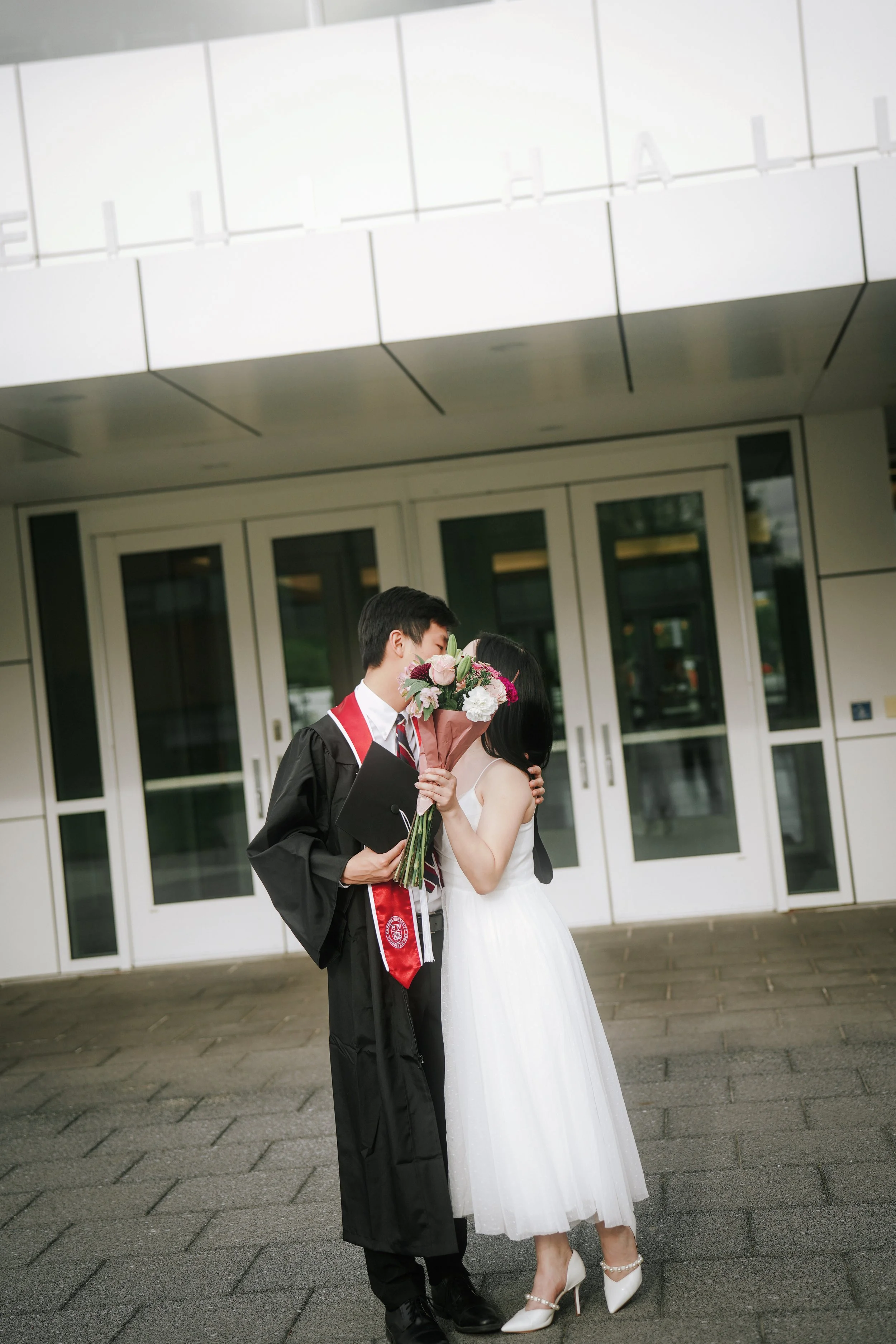 A young man in a graduation cap and gown holding a diploma and a bouquet standing close to a woman in a white wedding dress outside a building with glass doors.