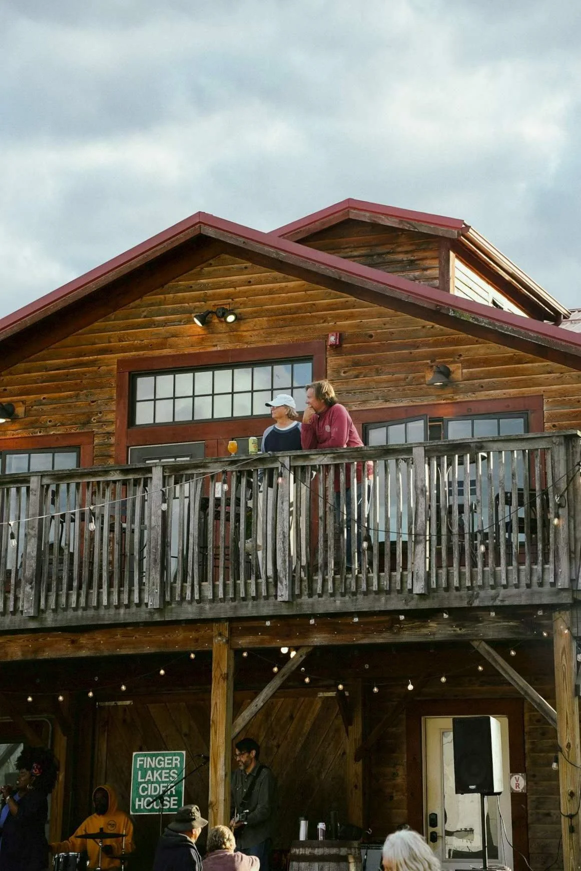People on a wooden balcony of a house, with a gathering below, including a band performing indoors and guests enjoying drinks outside.