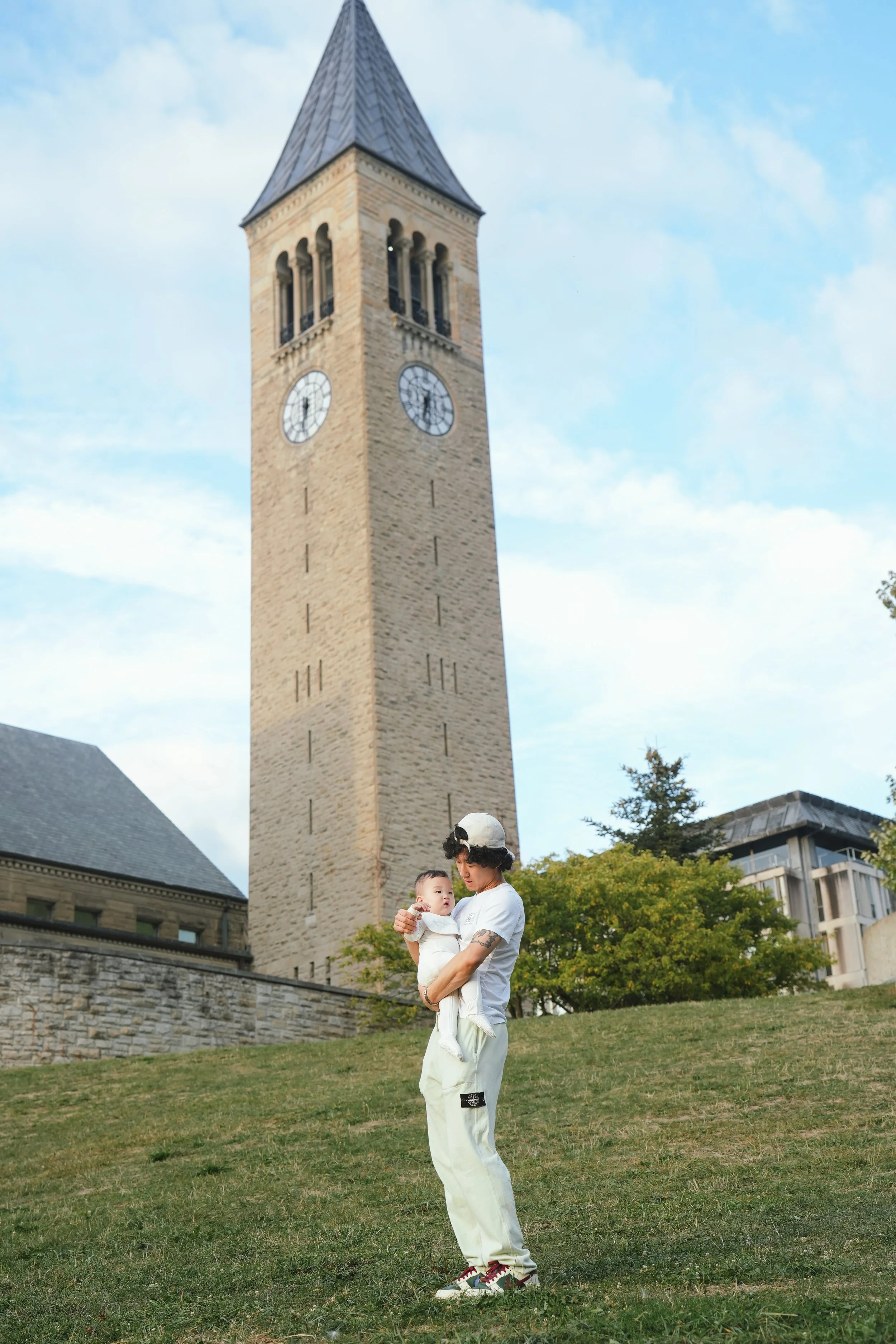 A man wearing a white hoodie and cream-colored pants holding a baby in his arms while standing on a grassy hill in front of a tall clock tower with a pointed metal roof, part of a historic building, under a cloudy sky. Trees and other buildings are v