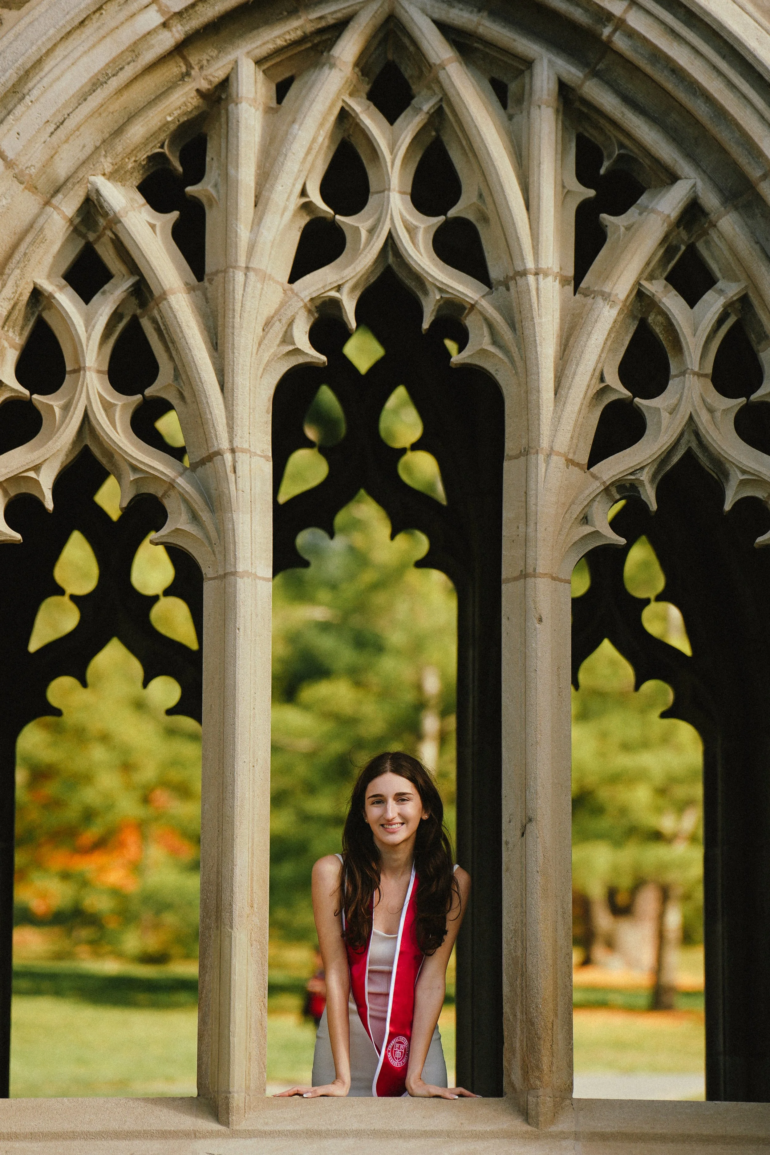 Young woman at graduation standing behind a stone architectural structure with Gothic arches, smiling.
