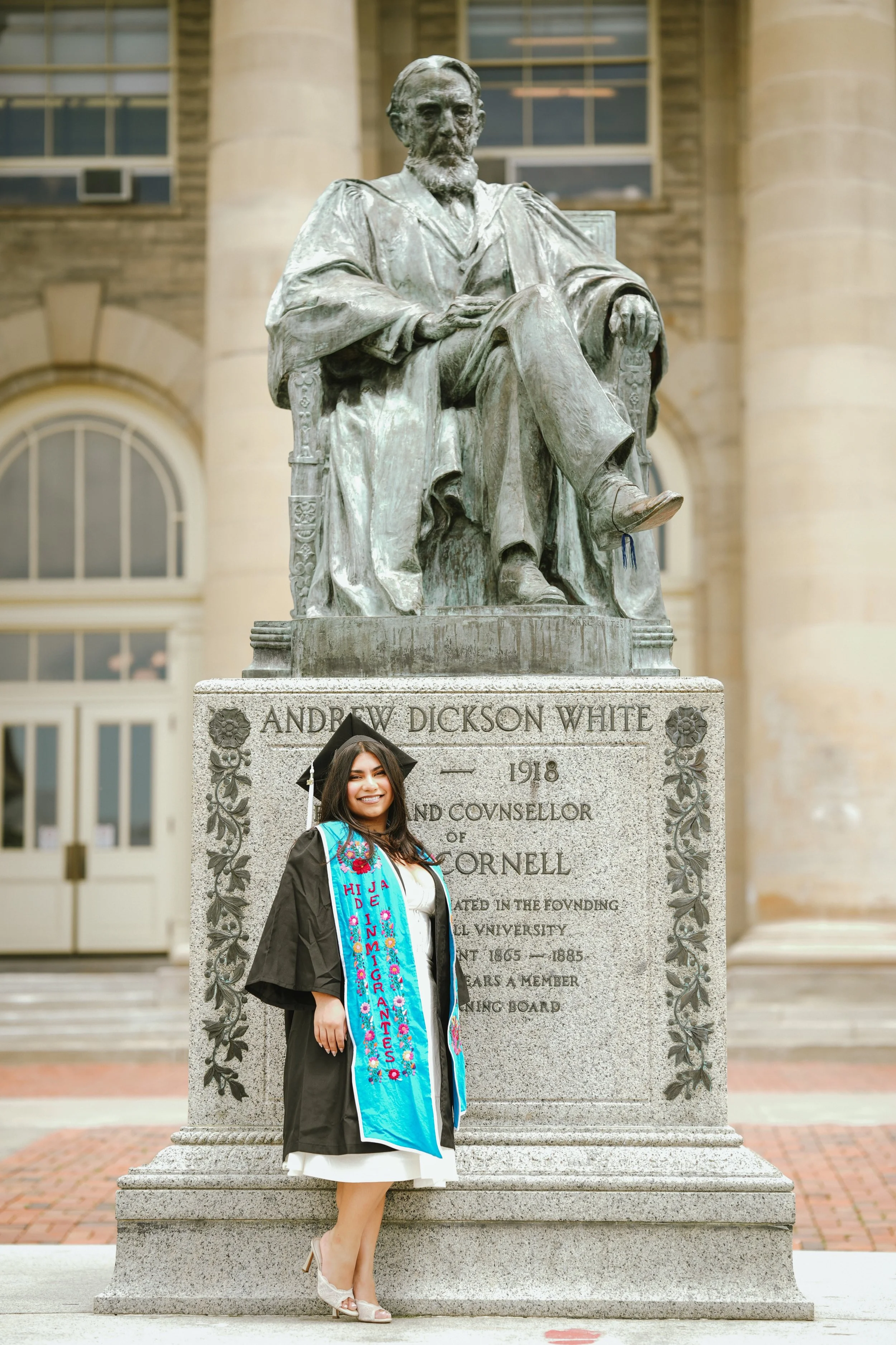 A young woman in graduation attire standing next to a large bronze bust statue of Andrew Dickson White, an academic figure, on a university campus. She is smiling and wearing a decorated graduation sash.