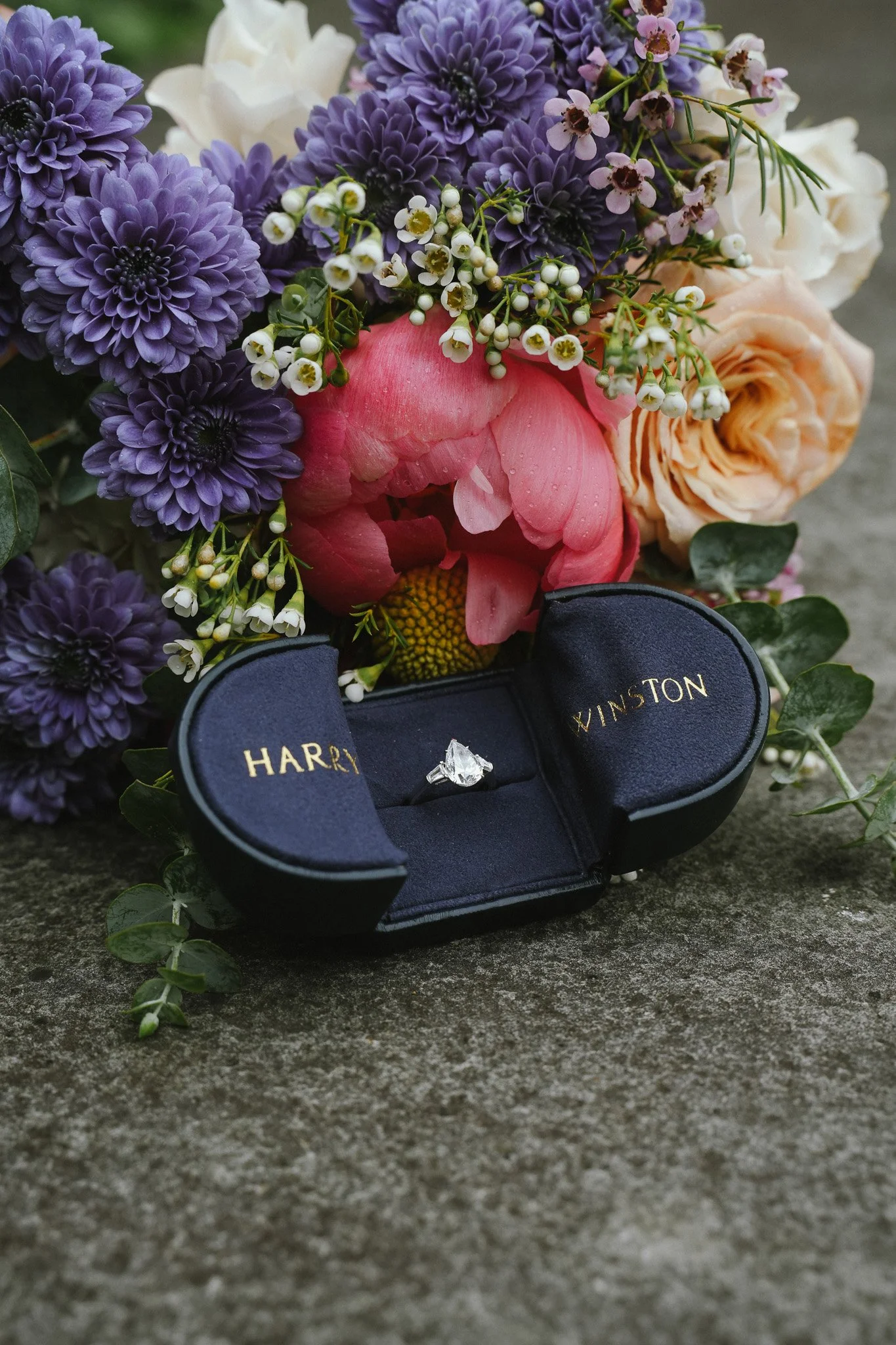 A bouquet of purple, white, pink, and peach flowers with an engagement ring in a blue velvet box resting on a concrete surface.