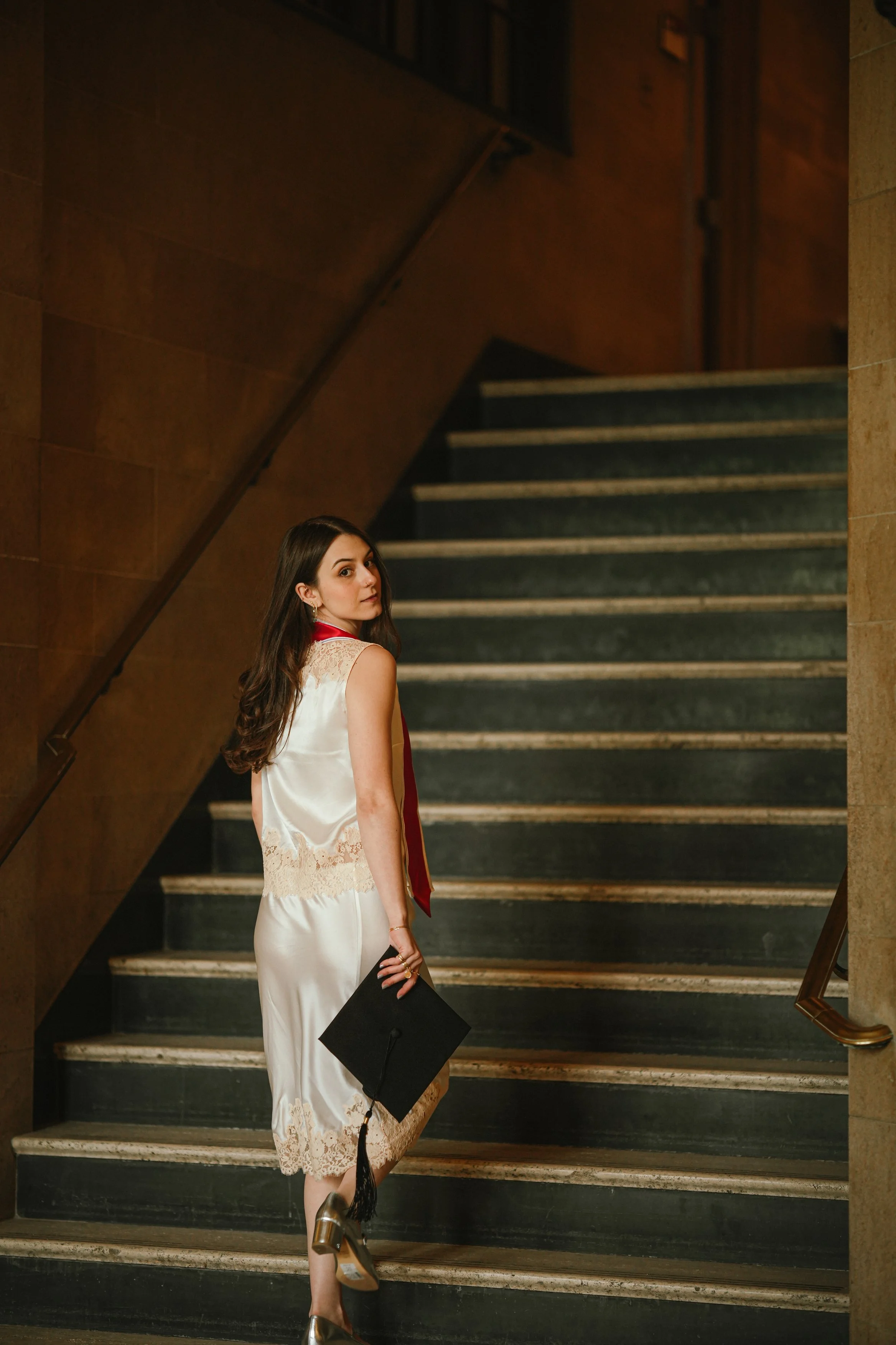 A young woman with long brown hair, wearing earrings and a white lace dress, stands on a staircase holding a black graduation cap in her right hand, looking over her shoulder.