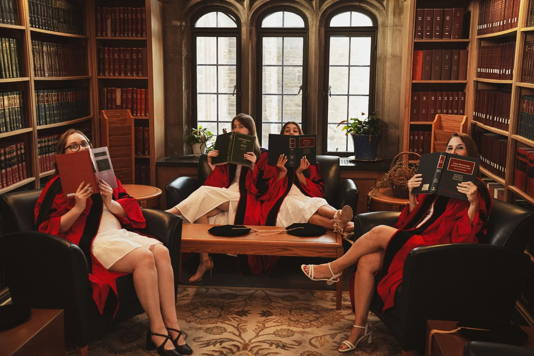 Four women dressed in graduation robes are sitting and lounging in a library room with tall bookshelves and large windows. They are holding books and smiling at the camera.