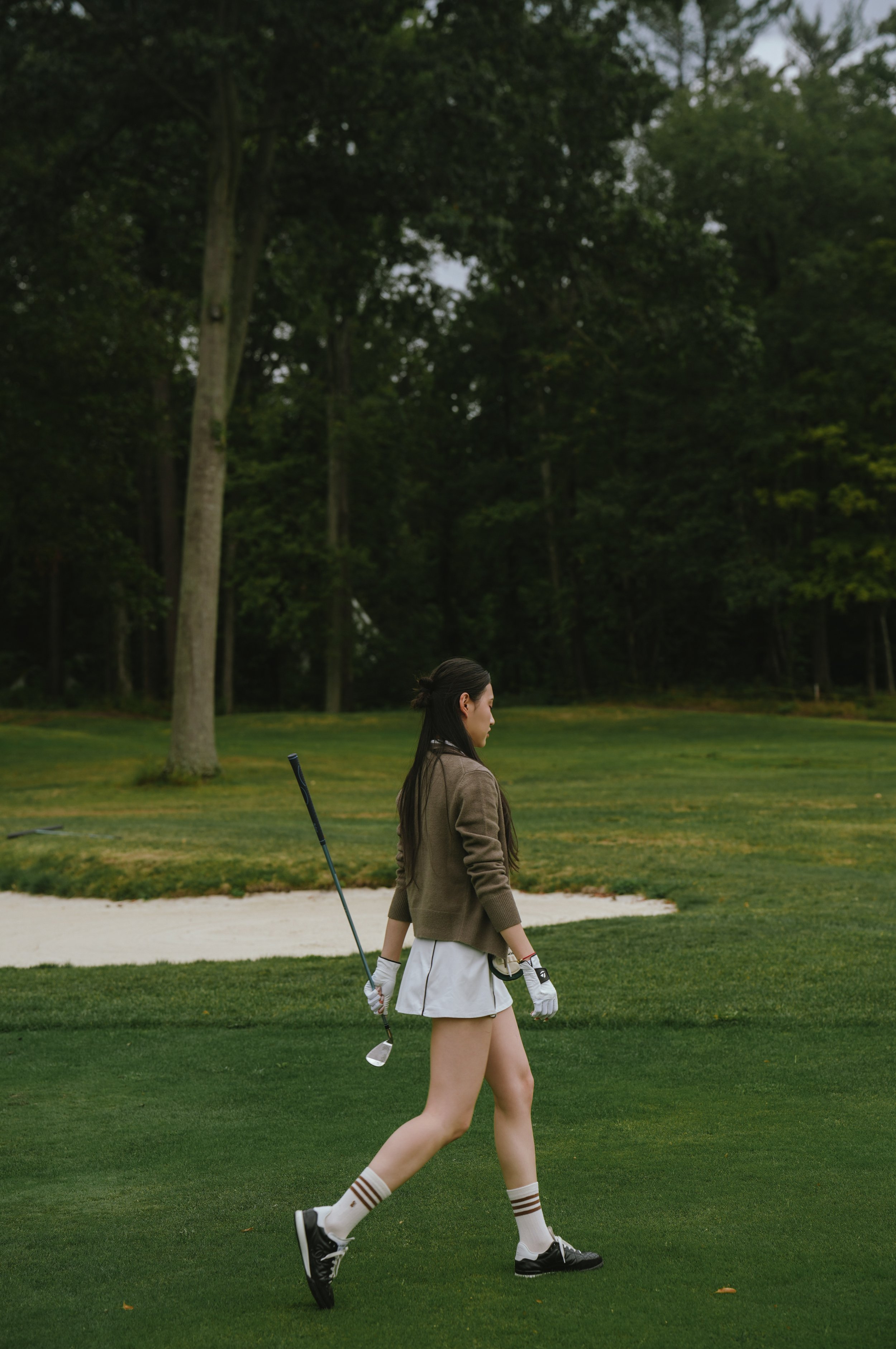A woman walking on a golf course wearing a brown jacket, white skirt, striped knee-high socks, and black golf shoes, carrying a golf club over her shoulder, with a lush green background and tall trees.