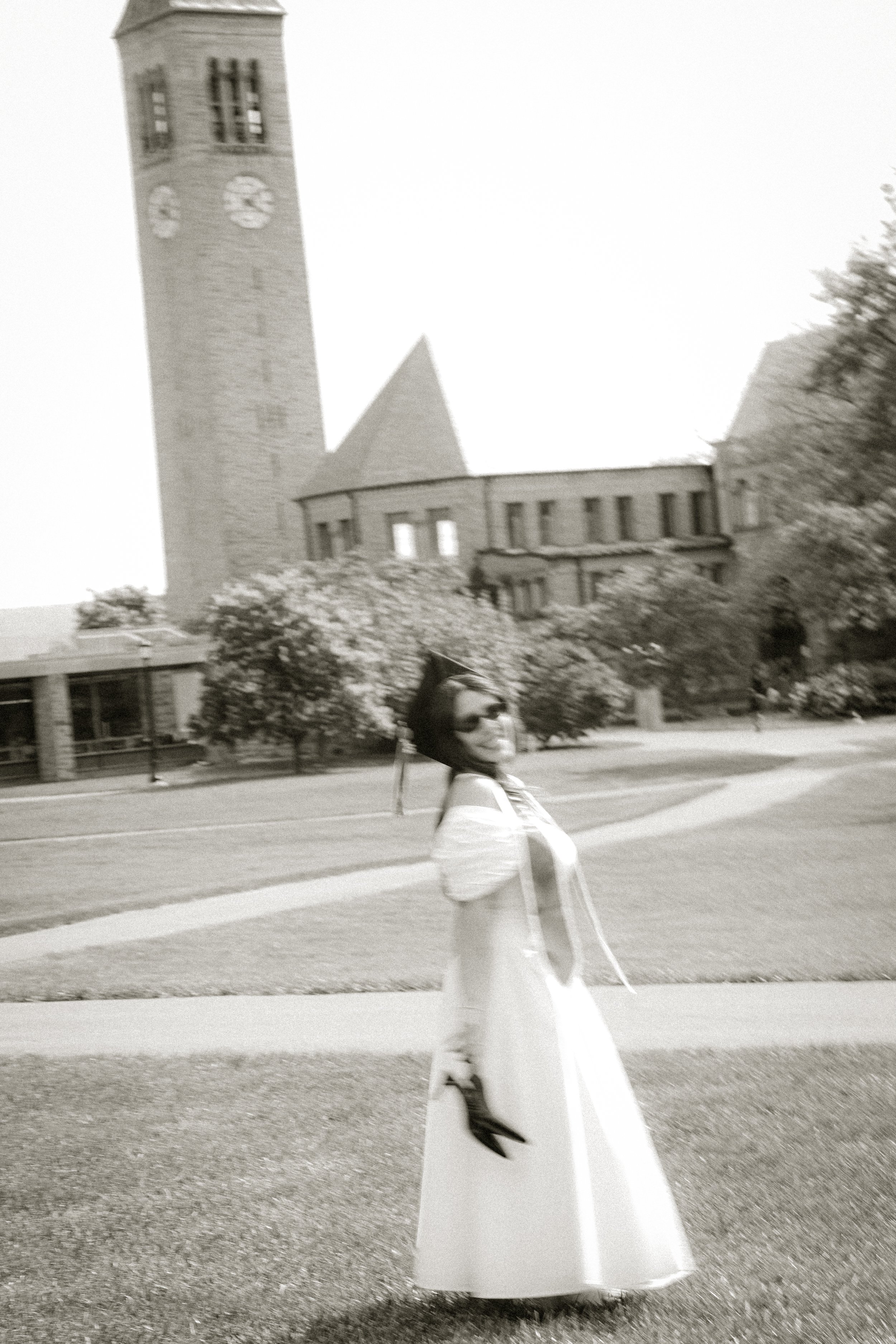 A woman wearing a long dress and sunglasses is walking on the sidewalk in front of a tall brick clock tower with trees and a building in the background.