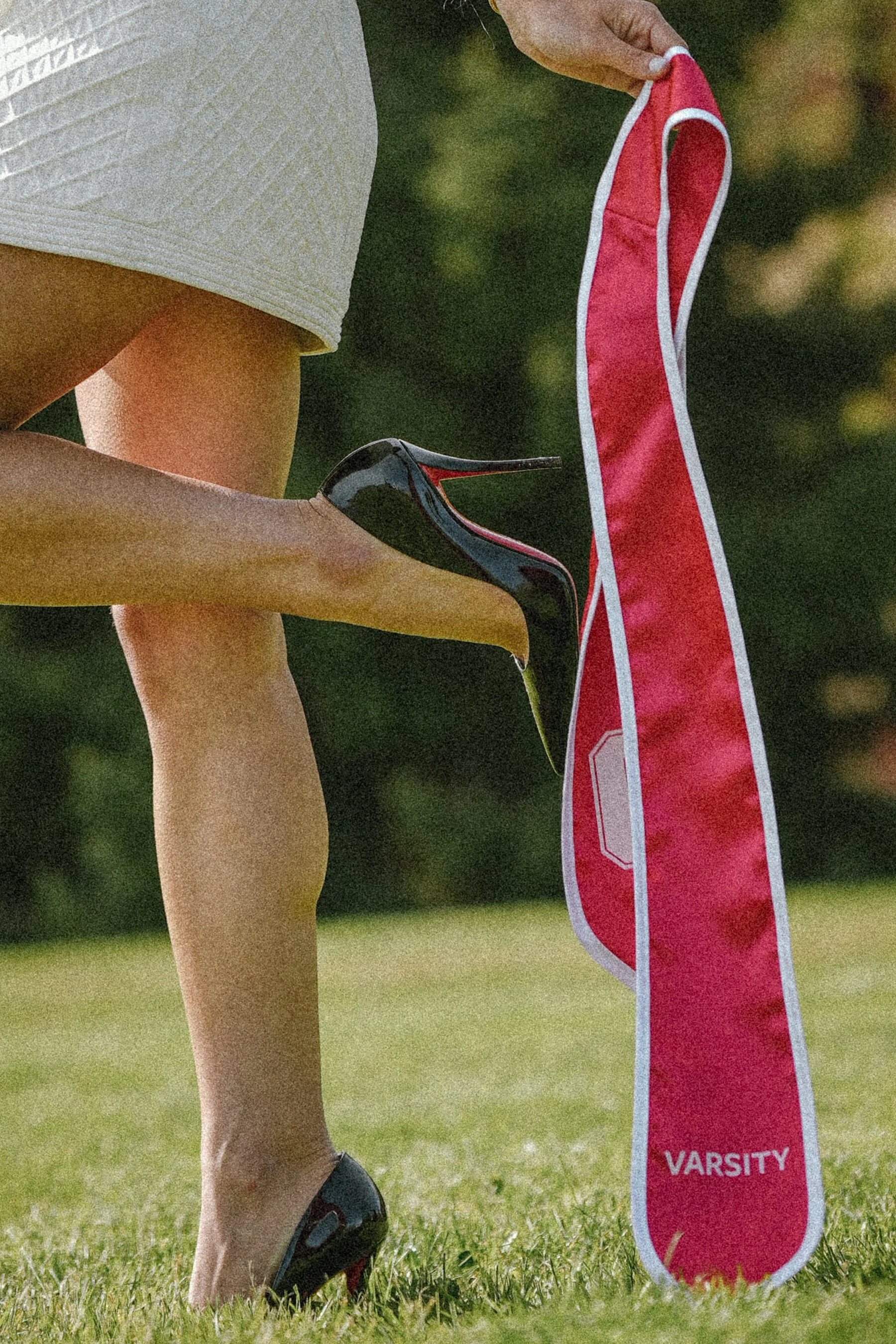 Close-up of a person’s legs, wearing black athletic shoes with red accents, standing on grass outdoors, next to a pink and white bag labeled 'VARSITY'.