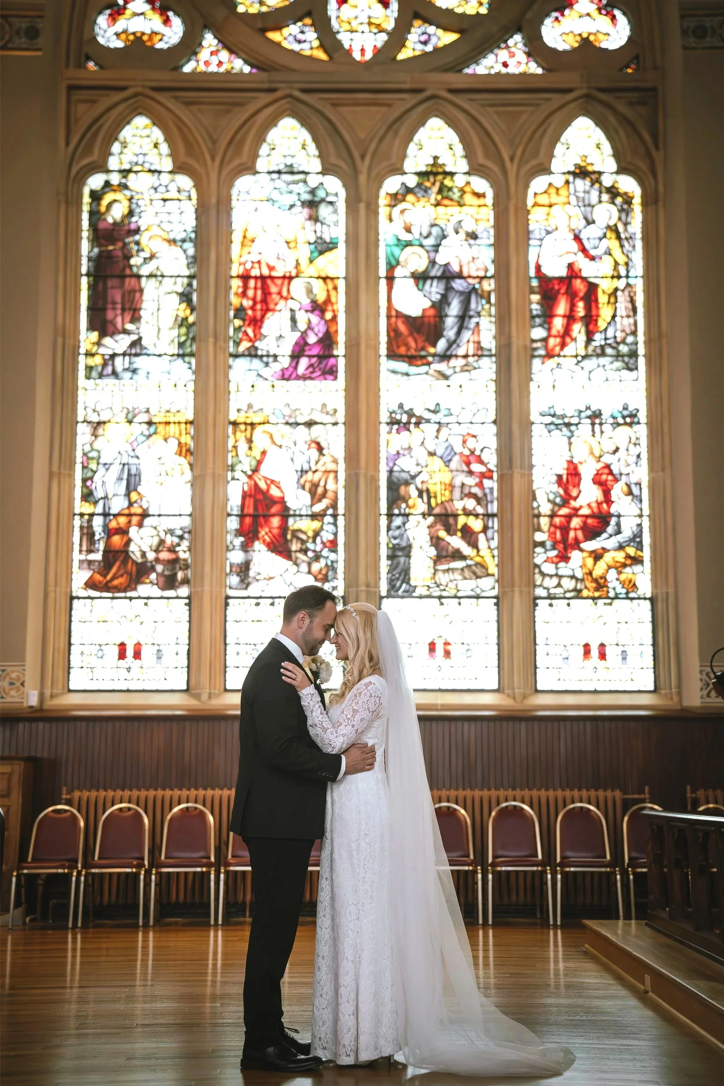 A bride and groom share a moment during their wedding inside a church with stained glass windows depicting biblical scenes.