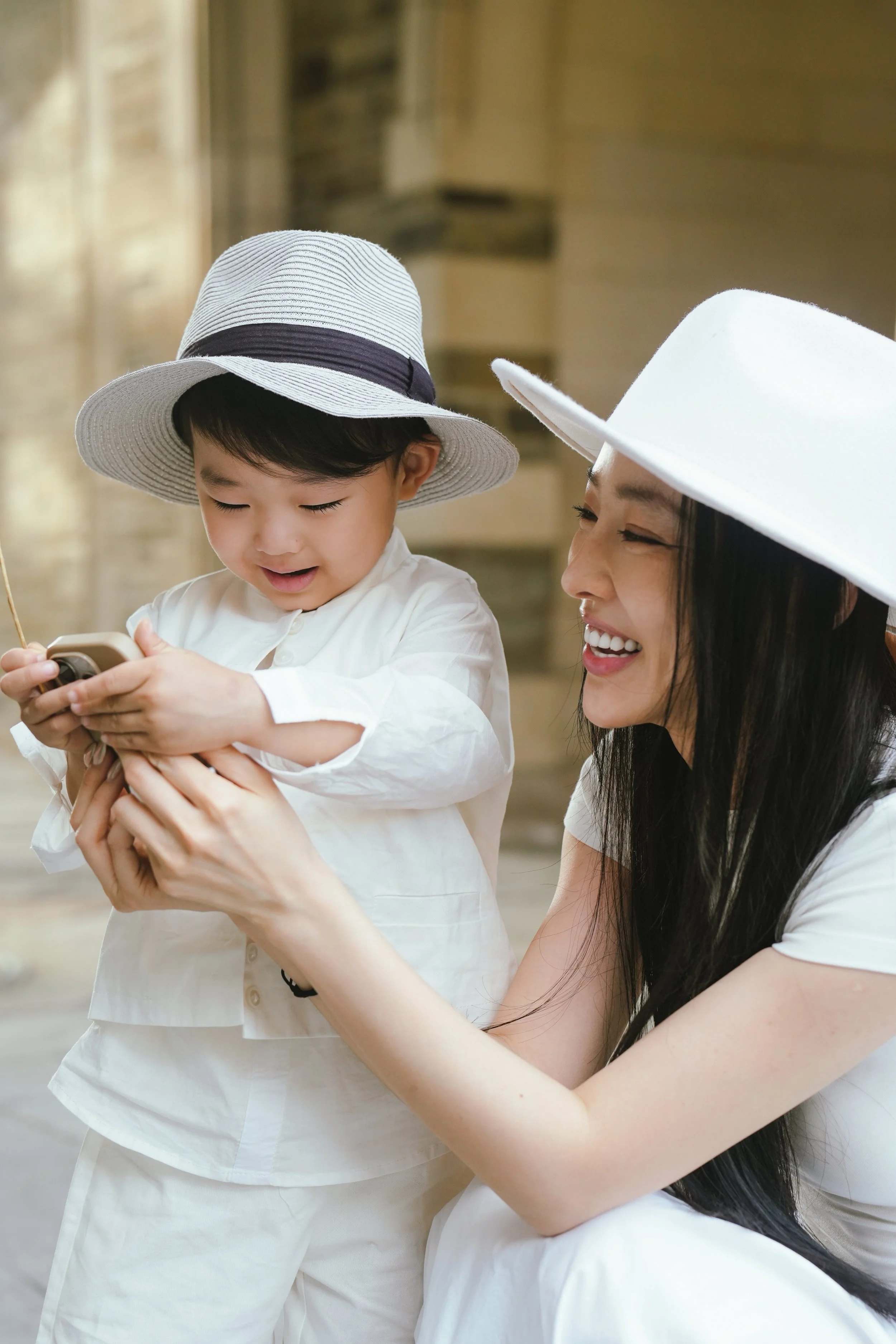 A woman with long black hair and a white wide-brimmed hat smiling at a little boy wearing a similar hat, both dressed in white, looking at a smartphone indoors.