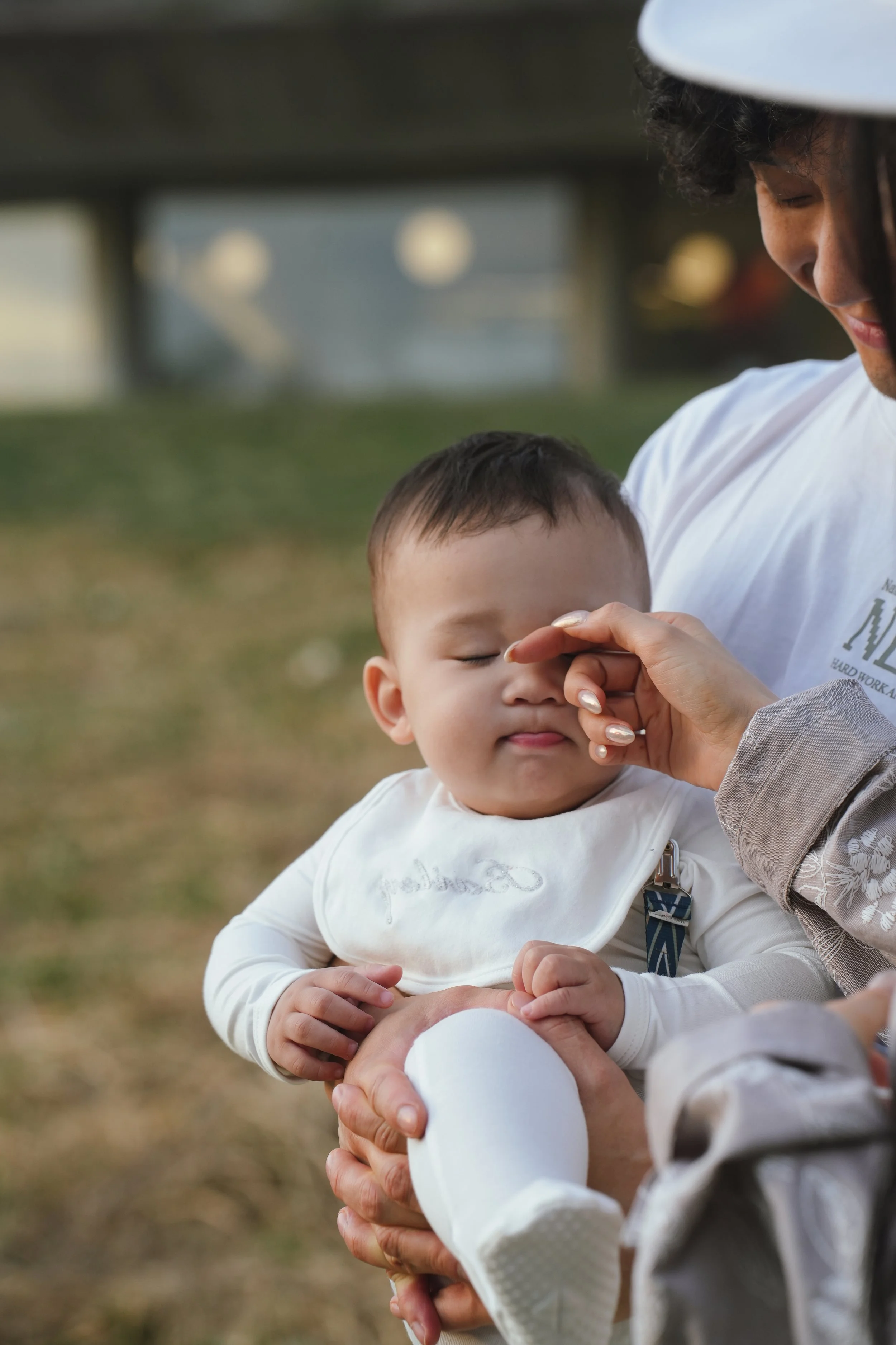 A man with curly hair wearing a white helmet is helping a child with short dark hair, who is sitting on his lap, with an activity outdoors in a blurred natural setting.