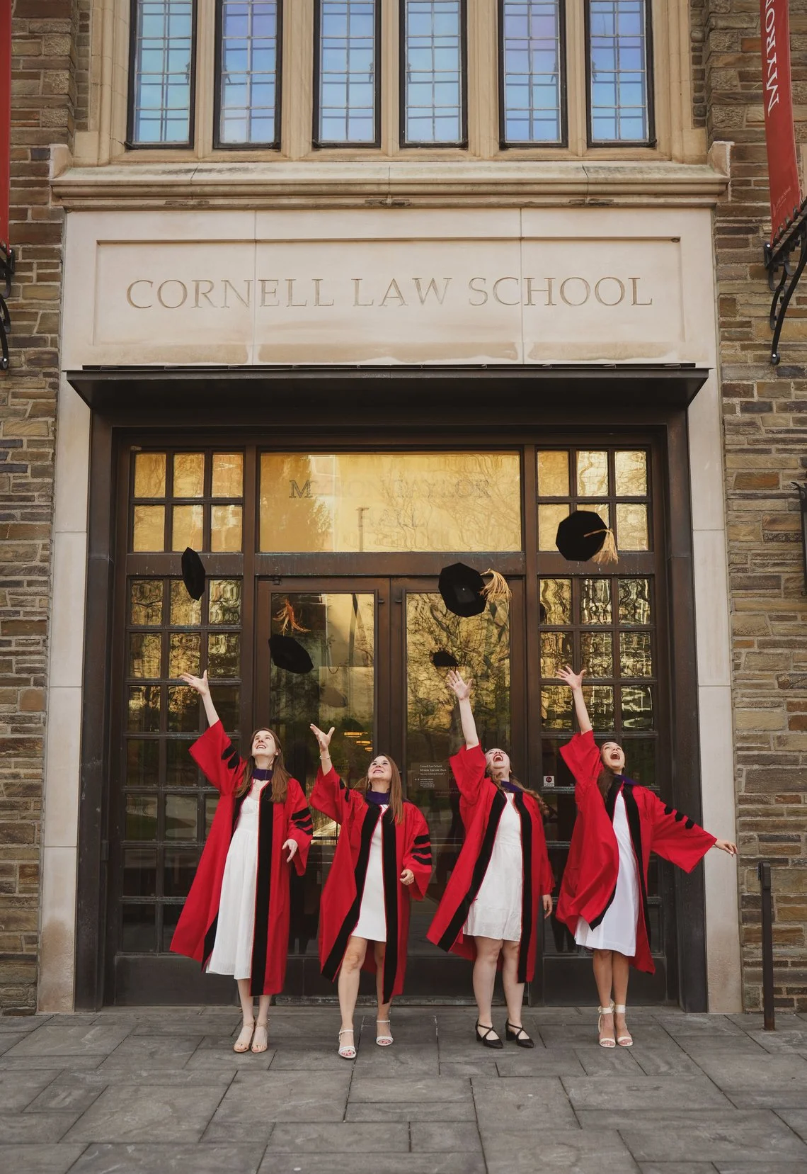 Four women in graduation gowns throwing caps in front of Cornell Law School building