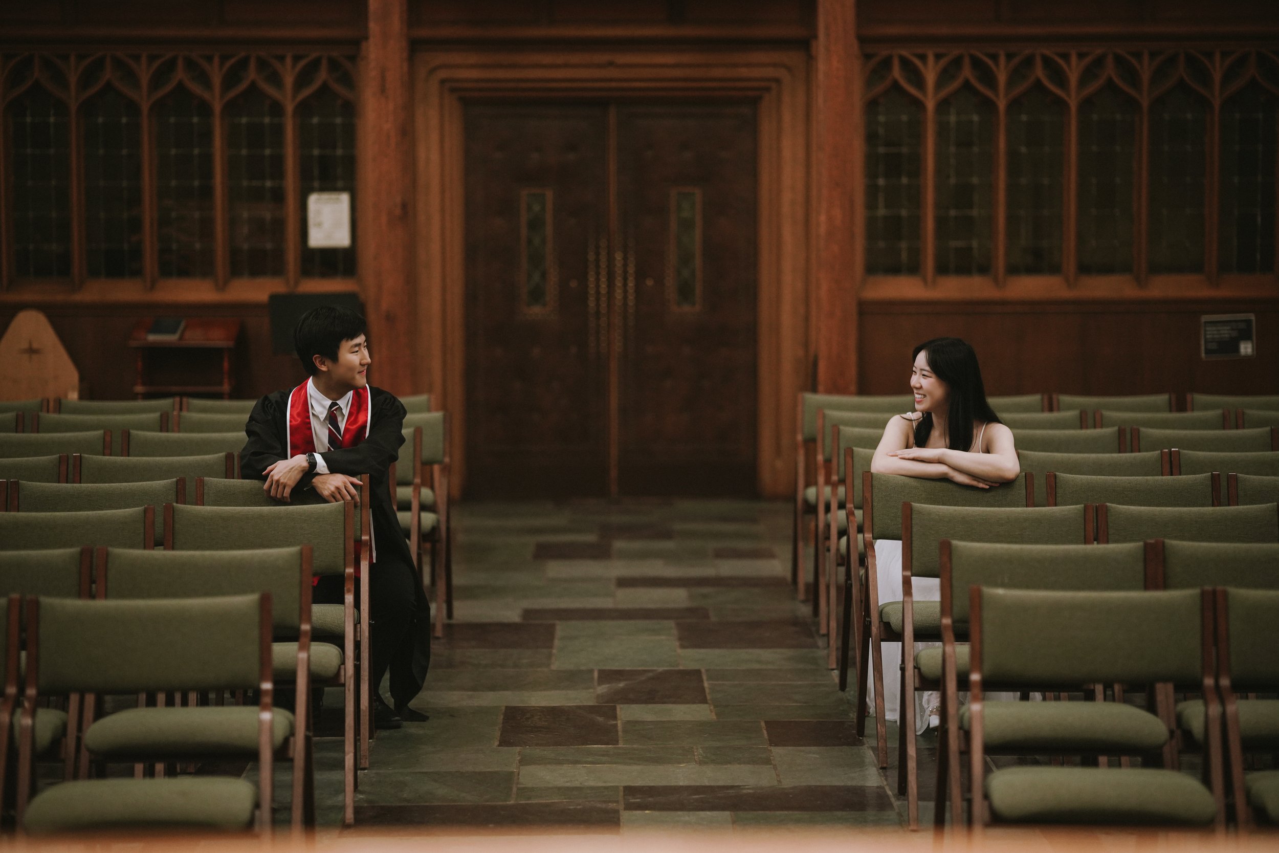 A young man in a graduation gown and tie sitting in an empty, wood-paneled hall, smiling at a young woman in a white dress sitting across from him.
