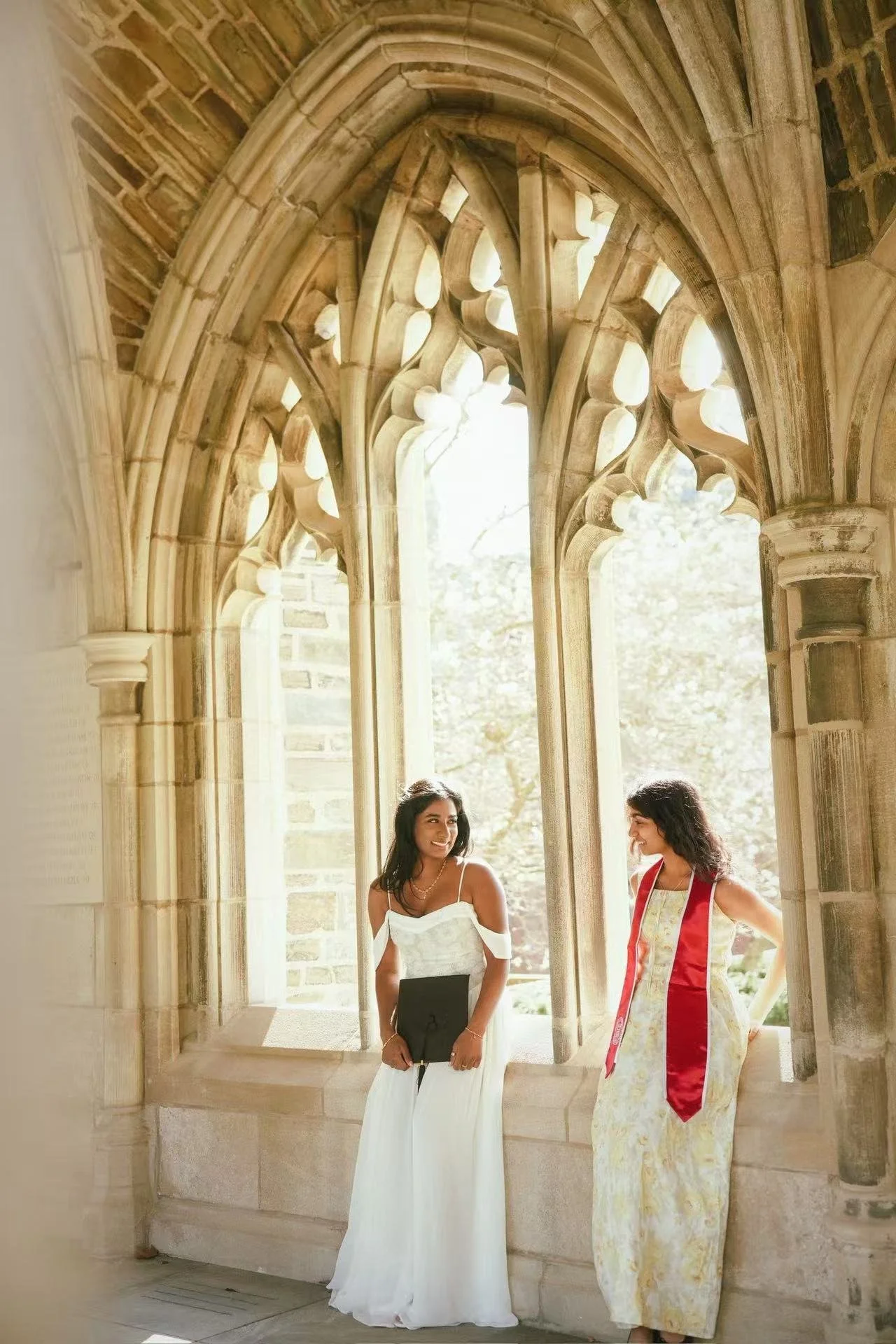 Two women in elegant dresses standing under a historic stone archway, smiling and engaging in conversation.