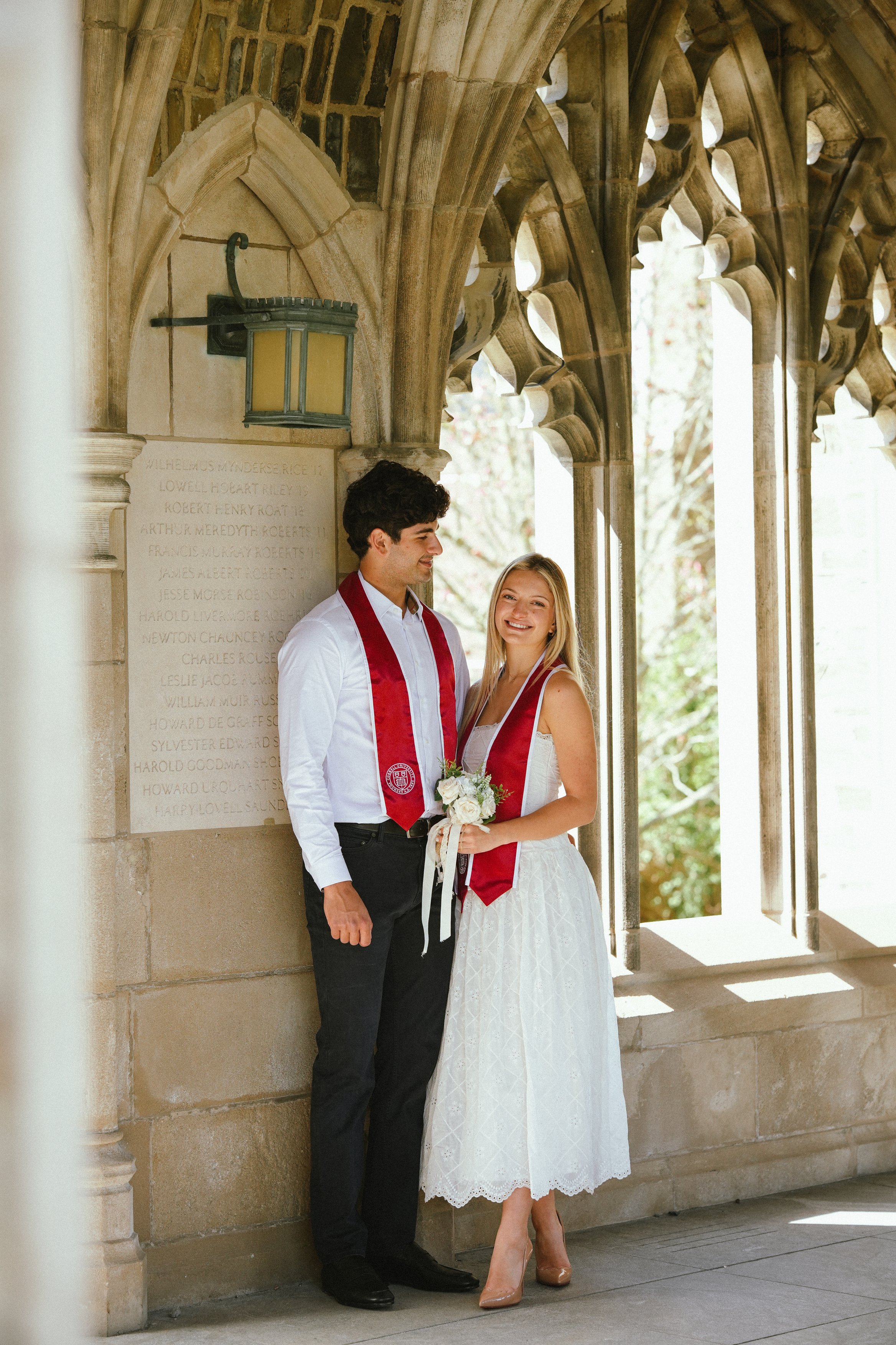 A young man and young woman standing together on a stone porch, dressed for a graduation ceremony, with the woman holding a bouquet of flowers. They are both wearing red graduation stoles and smiling.