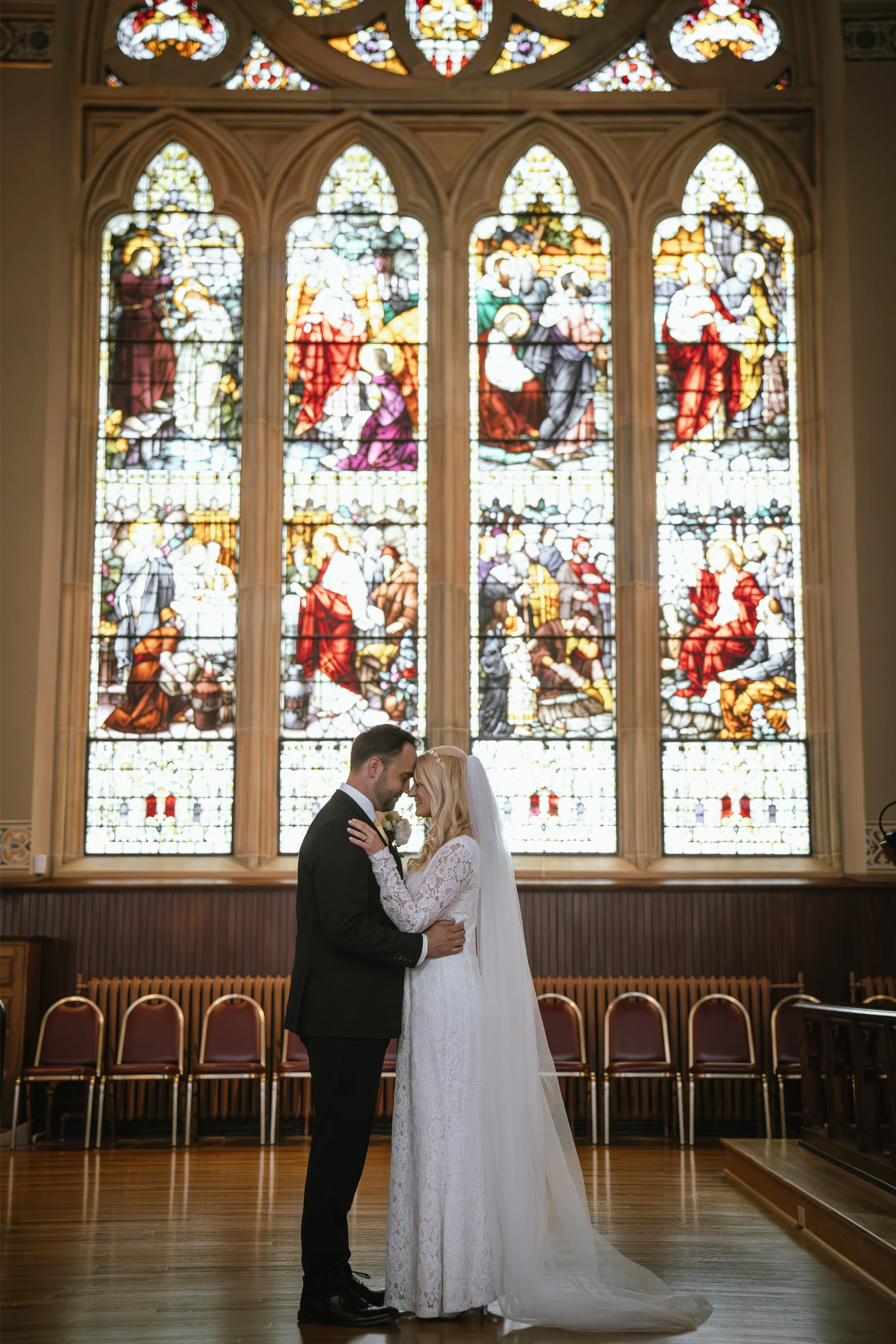 A bride and groom are standing close together in a church with stained glass windows behind them. They are touching foreheads and smiling, with the bride in a white lace wedding dress and veil, and the groom in a black suit and tie. Empty brown chairs are visible behind them.