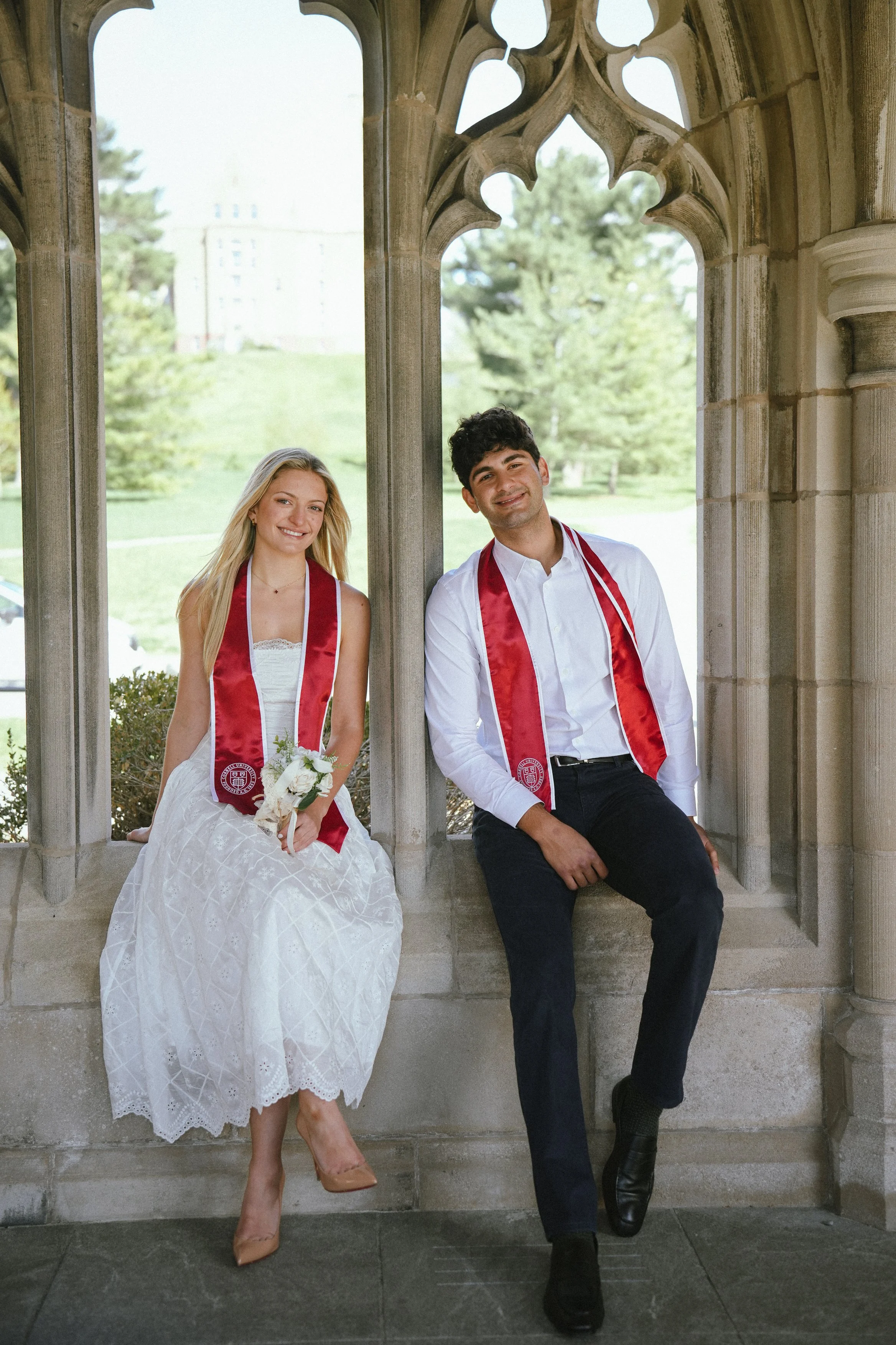A couple dressed in graduation attire, sitting on a stone ledge under an ornate stone archway, smiling at the camera with a background of green trees and a distant building.