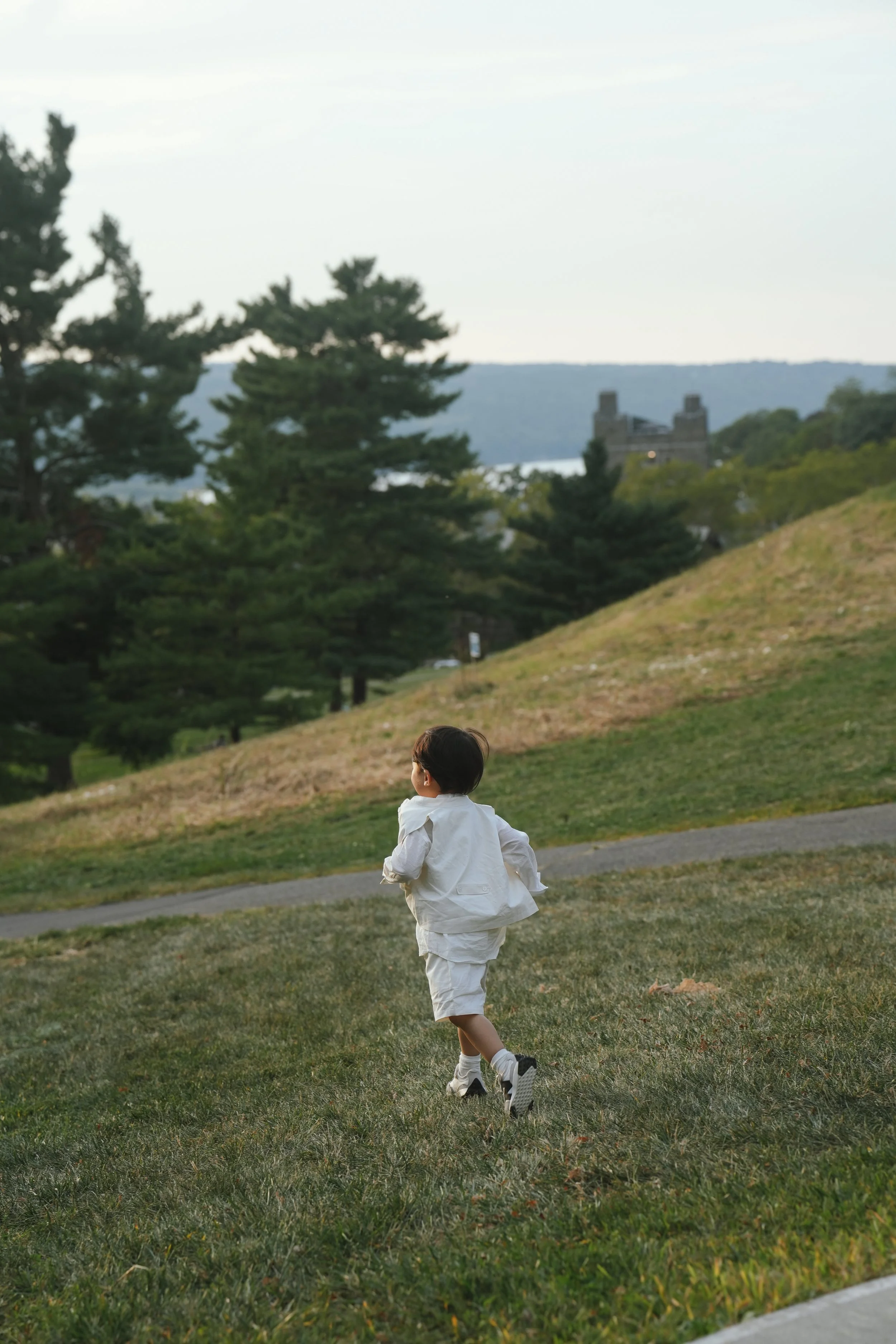 A young boy running on a grassy park at sunset, dressed in a white outfit with sneakers, with trees and a building visible in the background.