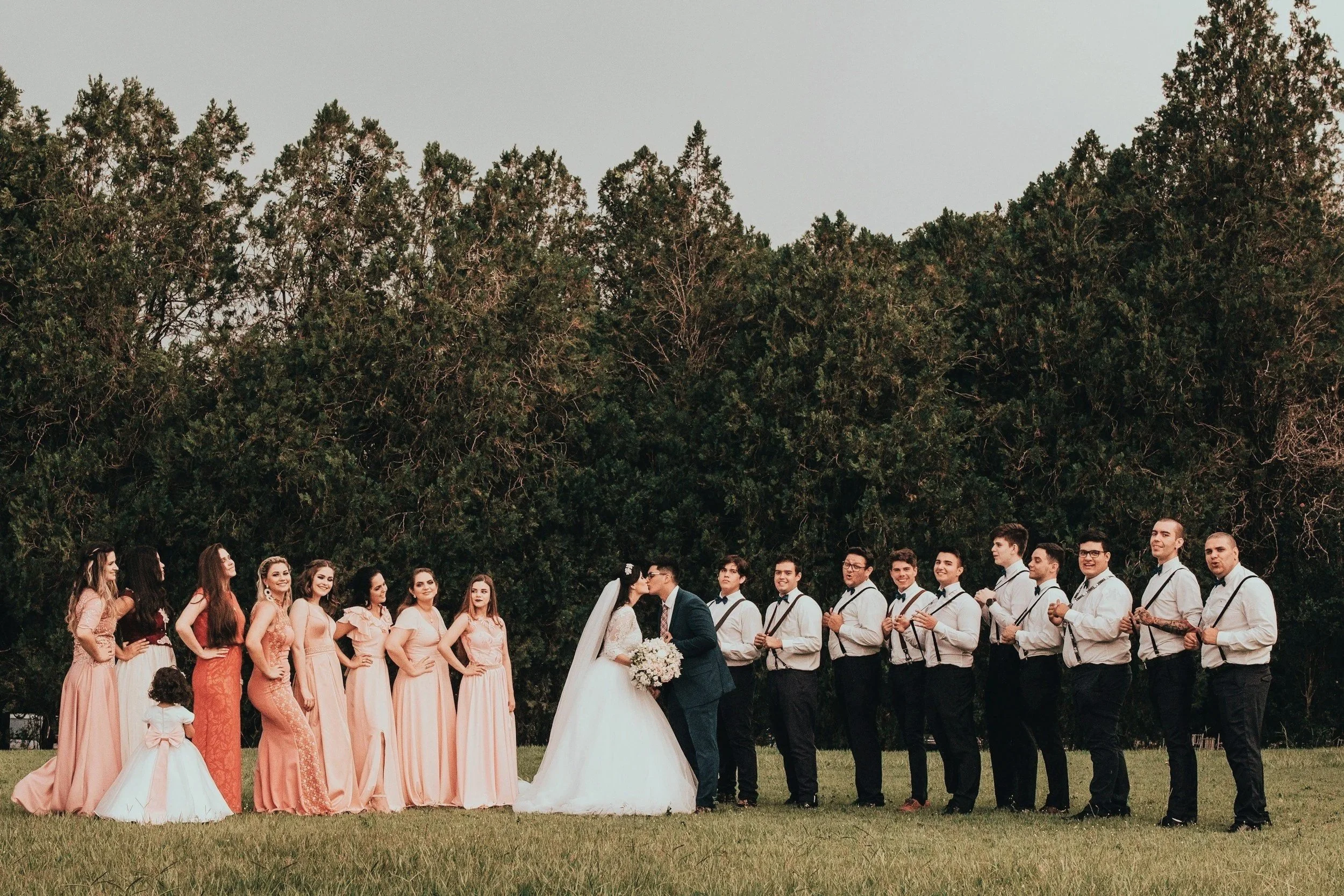 A large group of wedding guests standing outdoors on grass, posing for a photo in front of tall green trees. The bride and groom are kissing in the center, surrounded by bridesmaids in pink dresses and groomsmen in white shirts and suspenders.