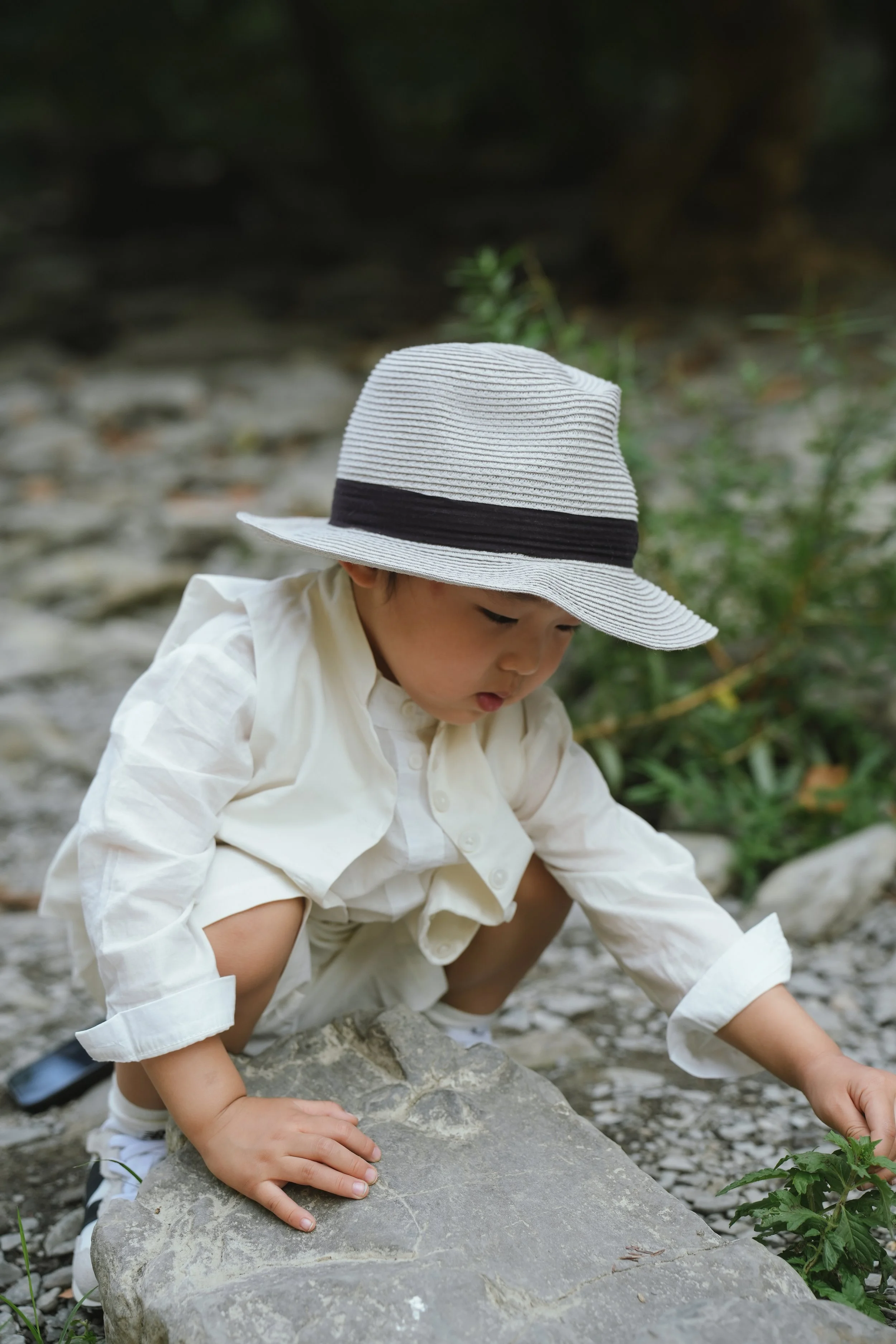 Young child wearing a white dress and wide-brimmed hat crouching near a rocky stream, exploring nature.