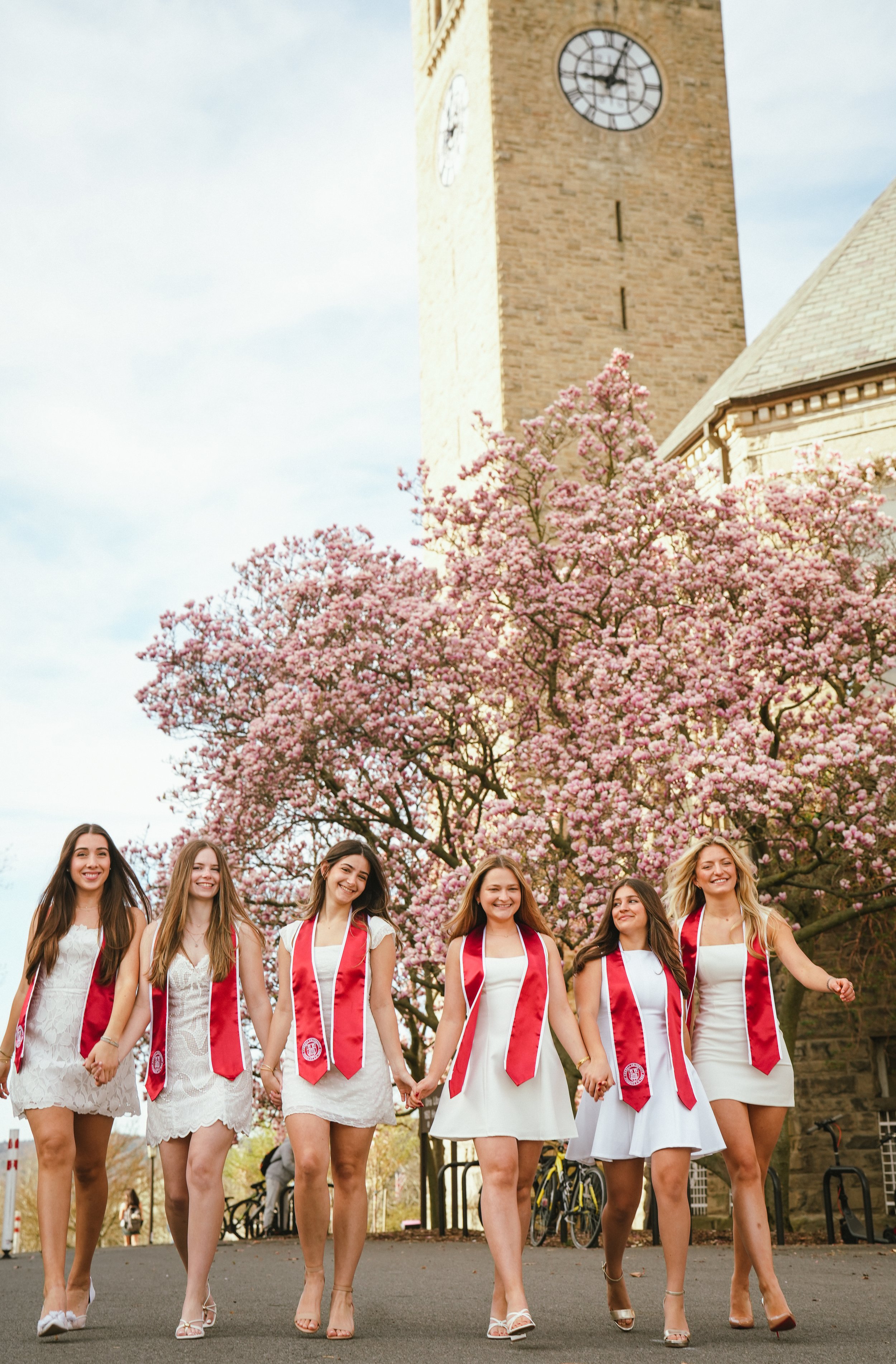 A group of six young women wearing white dresses and red sashes with college or university crests walk hand in hand outdoors during spring, with blooming pink cherry blossom trees and an old brick clock tower in the background.