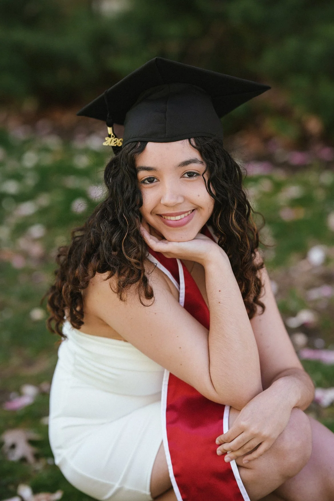 A young woman wearing a black graduation cap and a graduation gown with red and white accents, smiling and sitting outdoors with greenery and fallen flower petals in the background.
