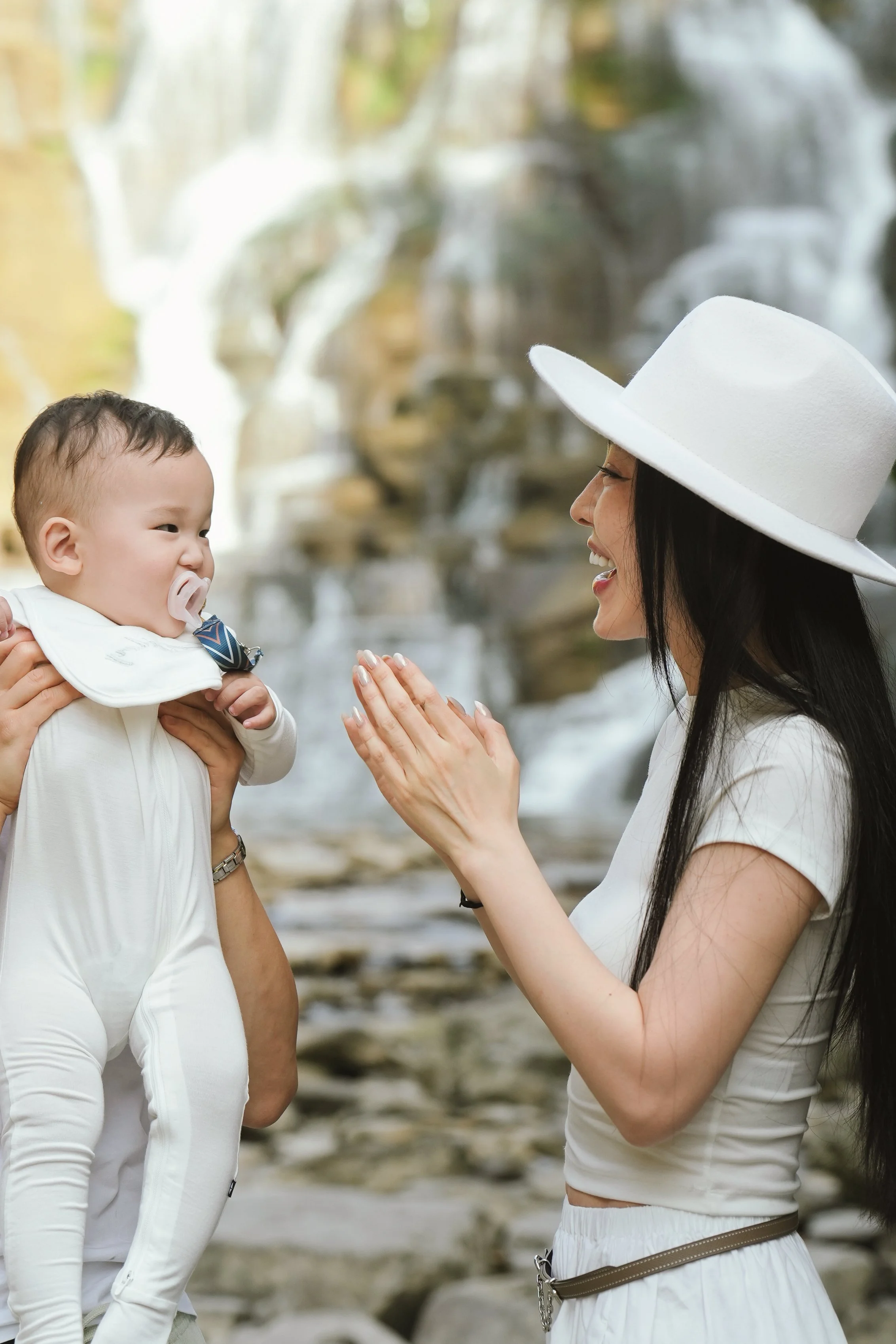 A woman wearing a wide-brimmed white hat is smiling and talking to a baby, who is being held up by an adult. The baby has a pacifier in their mouth and is dressed in white with a navy blue bow tie. The background features a blurred waterfall and rock