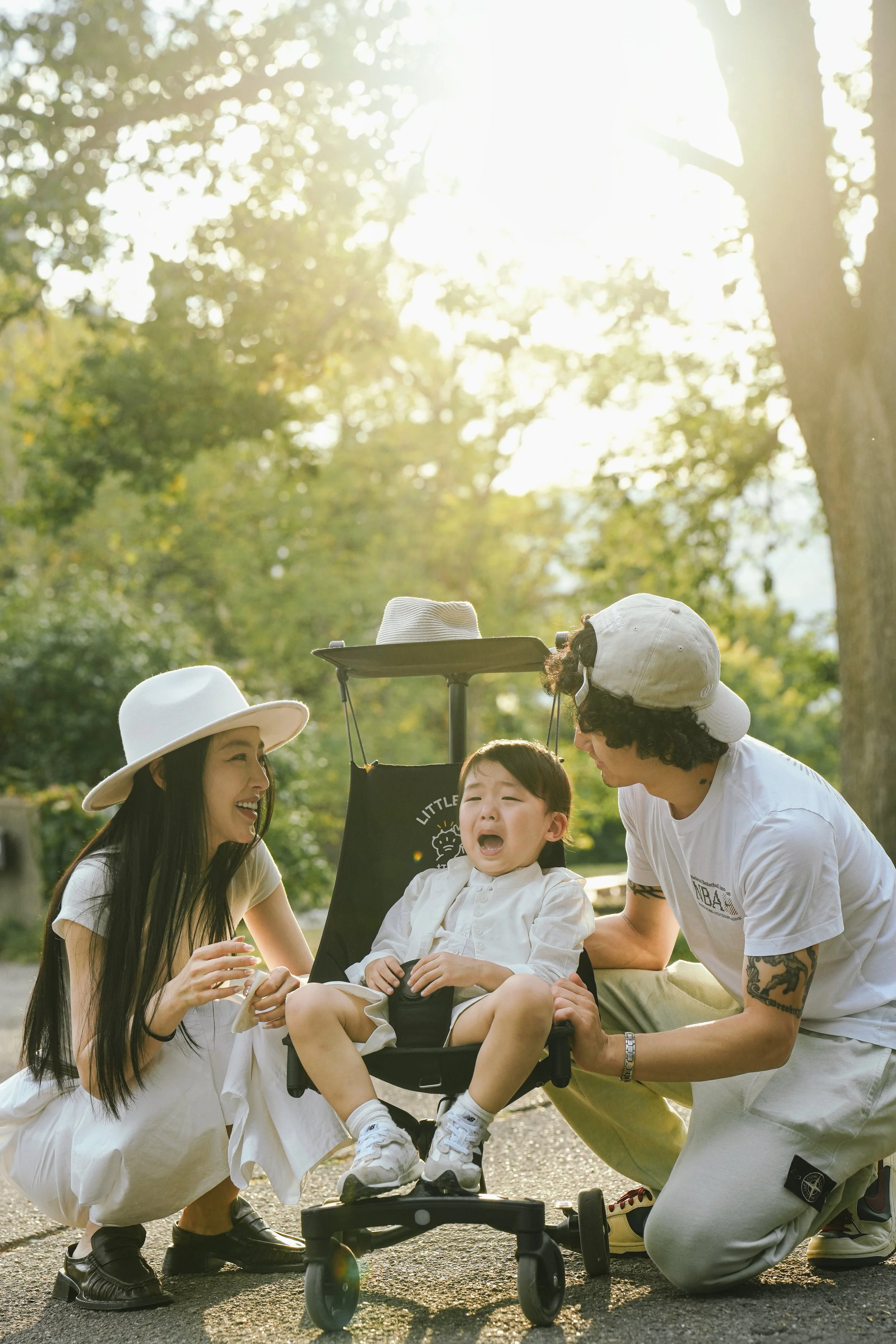 A young boy with a distressed expression sitting in a stroller, surrounded by three adults with diverse appearances, in an outdoor setting with trees and sunlight.